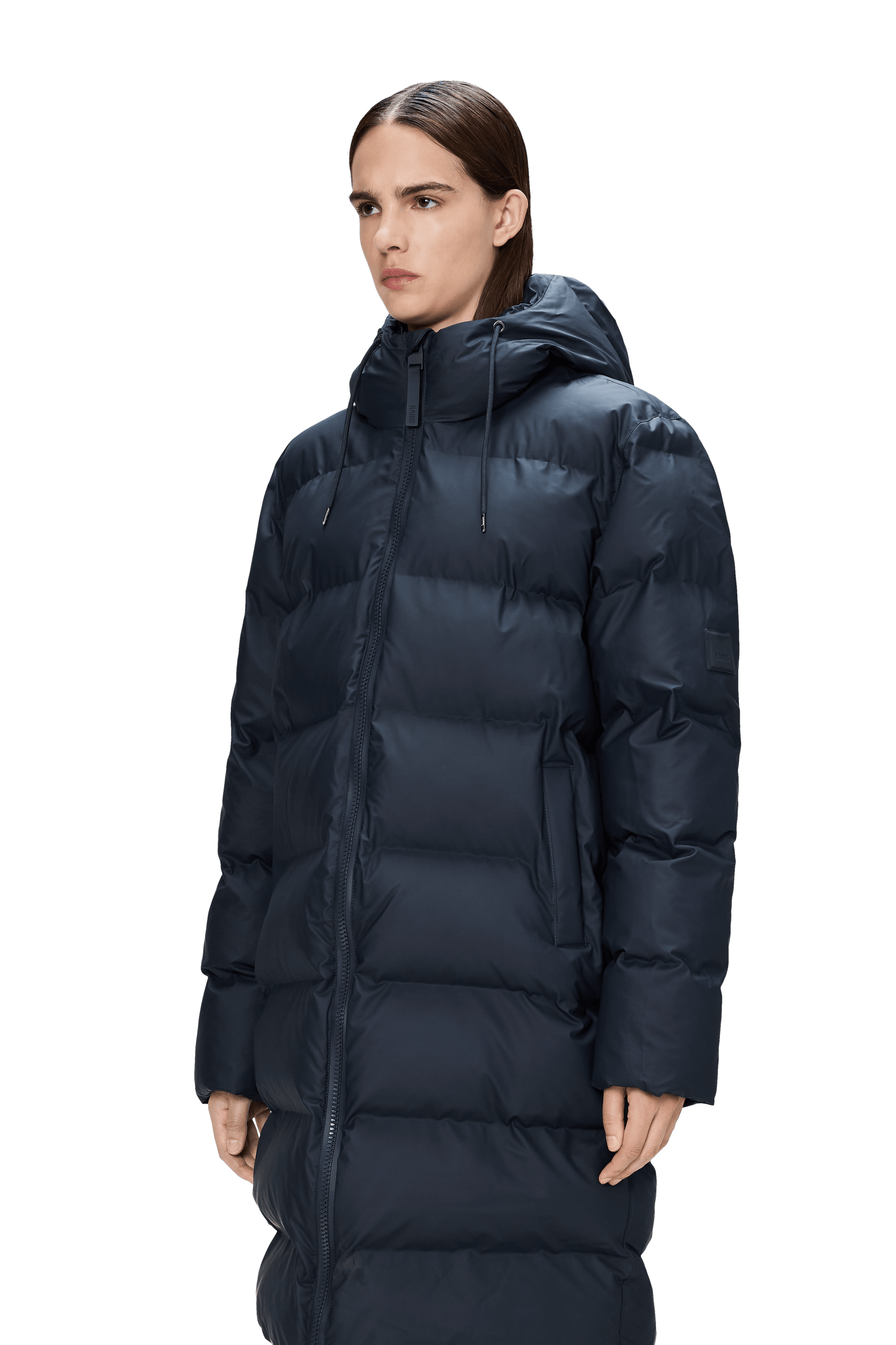 A person wearing the Rains Alta Puffer Longer in Navy—a hooded, featherless insulated winter jacket—stands against a plain light background.