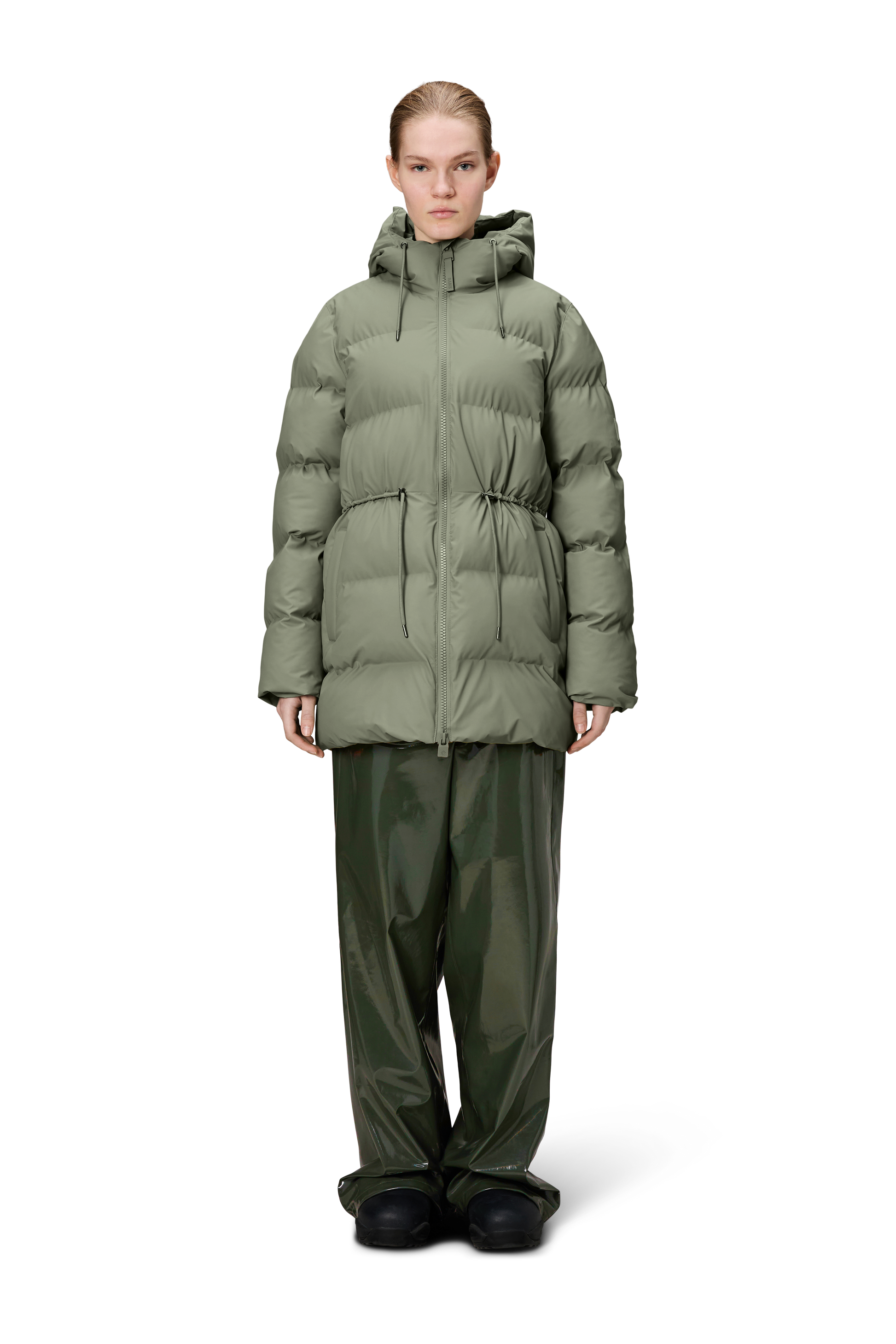 A person stands facing forward against a plain white background, wearing the Rains Alta Long Puffer String Jacket - Drift (olive green) with wide-leg green pants and black shoes.