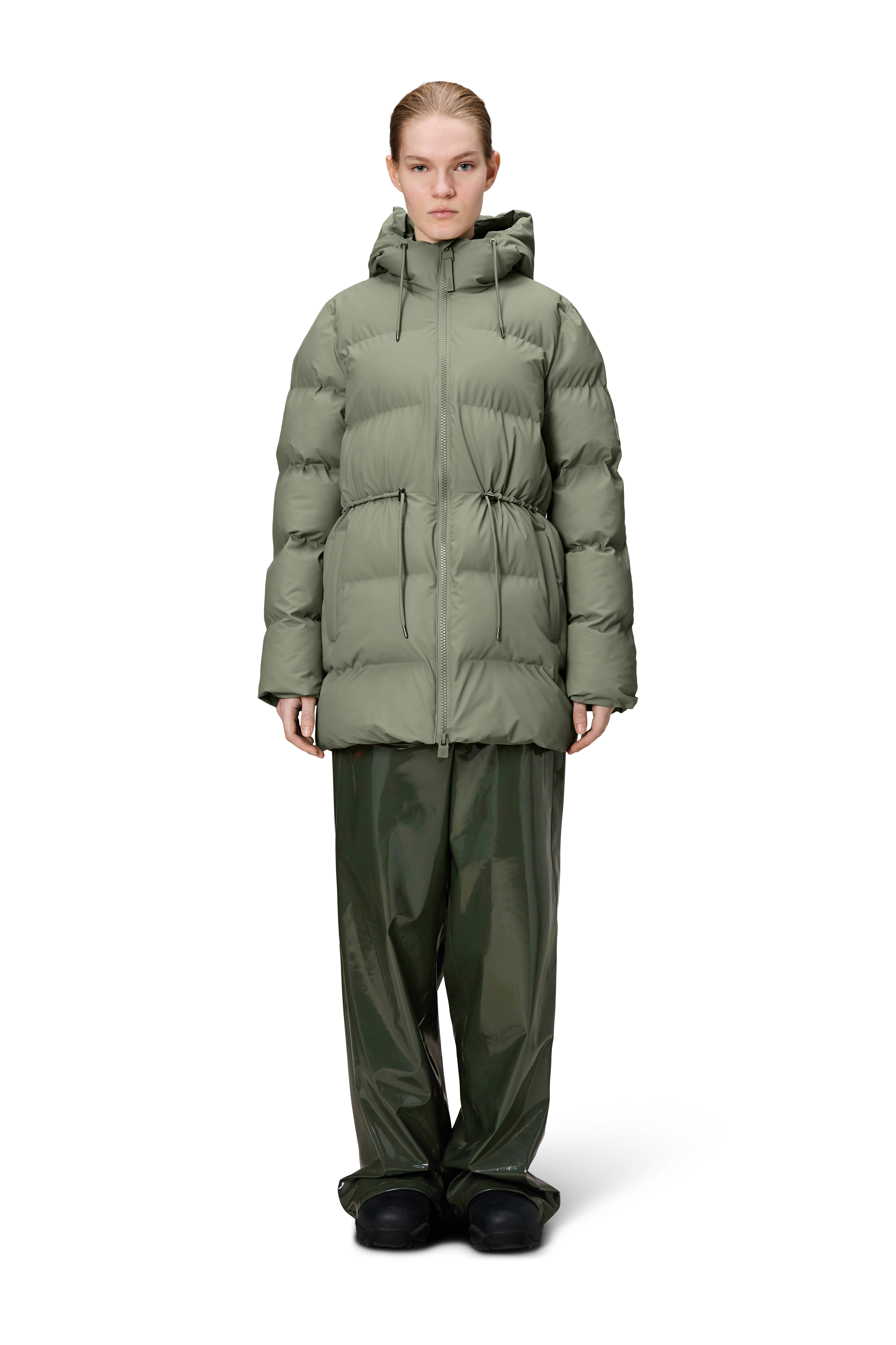 A person stands facing forward against a plain white background, wearing the Rains Alta Long Puffer String Jacket - Drift (olive green) with wide-leg green pants and black shoes.