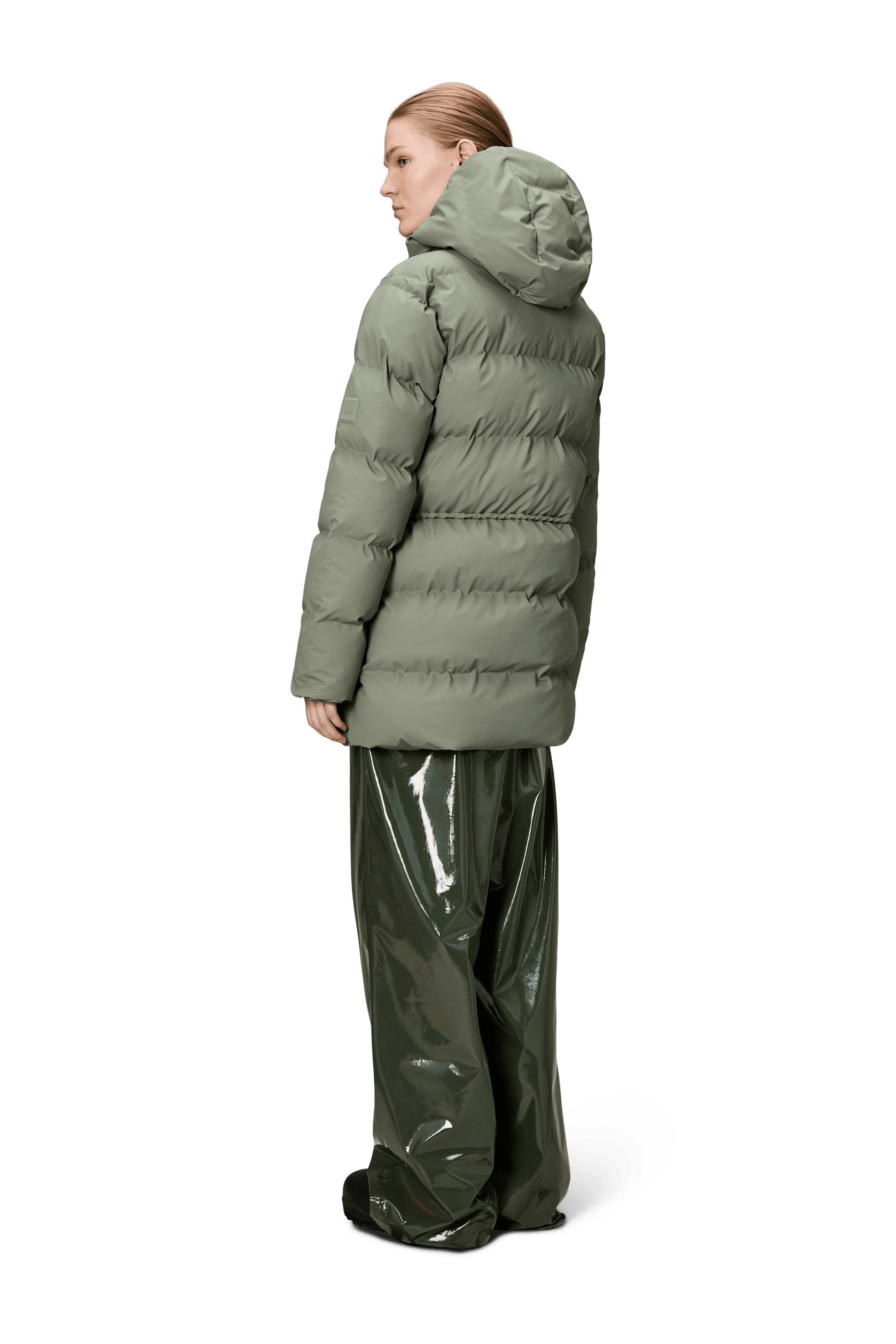 A person faces away, wearing the Rains Alta Long Puffer String Jacket in Drift with an insulated hood, paired with shiny, loose dark green pants, all set against a plain white background.