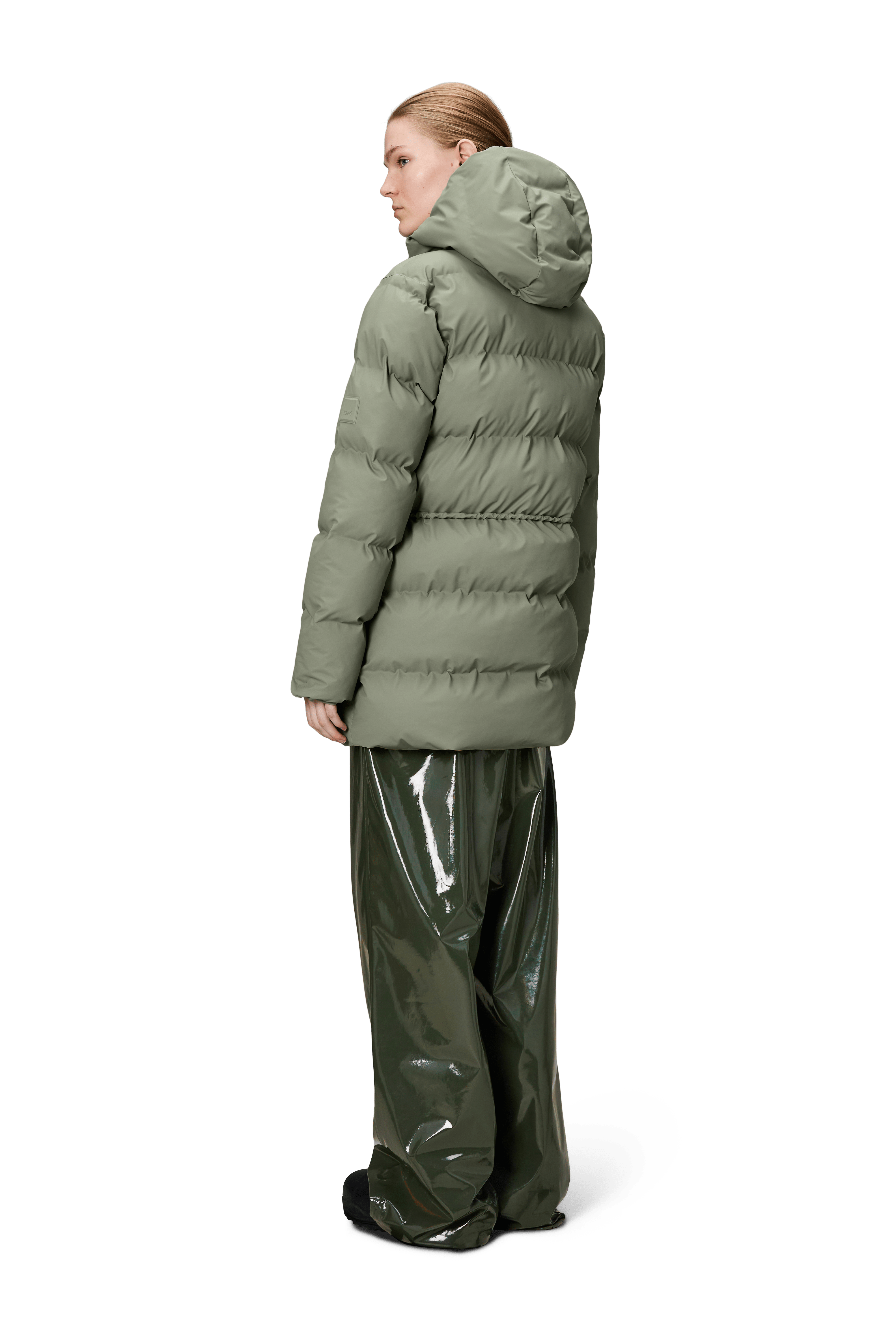 A person faces away, wearing the Rains Alta Long Puffer String Jacket in Drift with an insulated hood, paired with shiny, loose dark green pants, all set against a plain white background.
