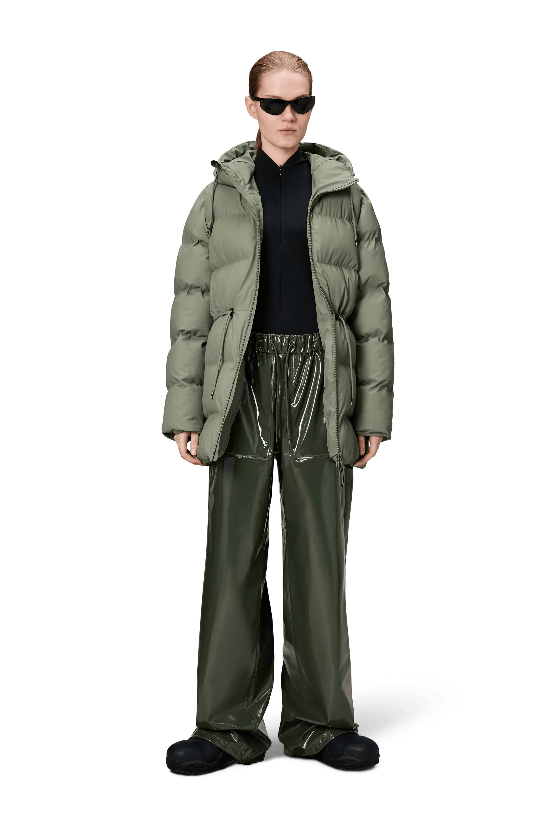 A person wears the Rains Alta Long Puffer String Jacket in Drift, paired with a black top, shiny green wide-leg pants, black shoes, and sunglasses, standing against a plain white background.