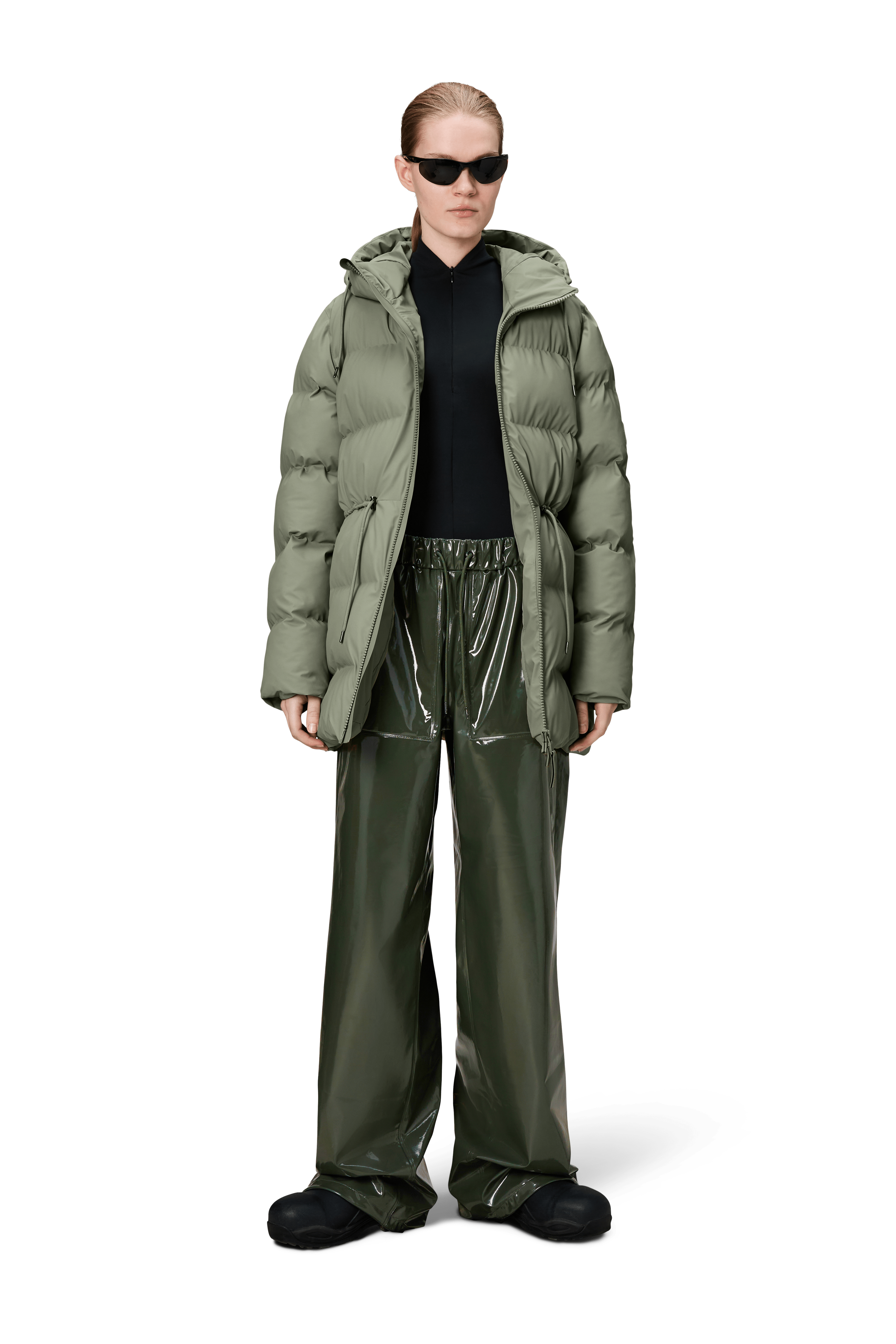 A person wears the Rains Alta Long Puffer String Jacket in Drift, paired with a black top, shiny green wide-leg pants, black shoes, and sunglasses, standing against a plain white background.