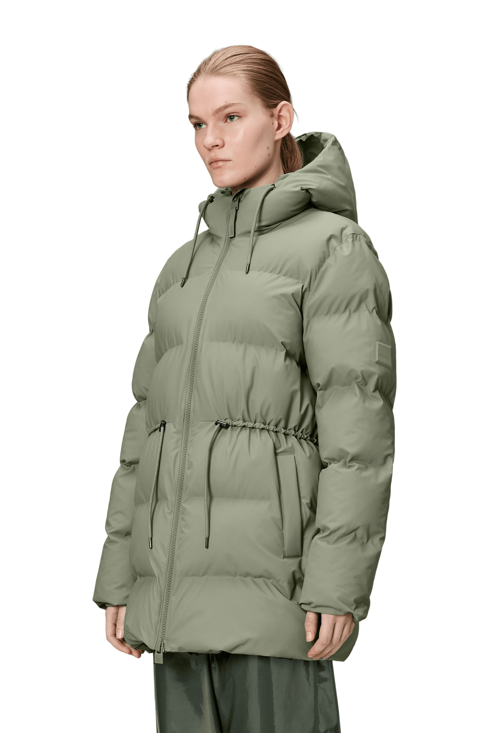 A person wearing the Alta Long Puffer String Jacket - Drift by Rains, a light olive green waterproof winter coat with an insulated hood, front zipper, drawstring waist, and side pockets, stands against a plain background.