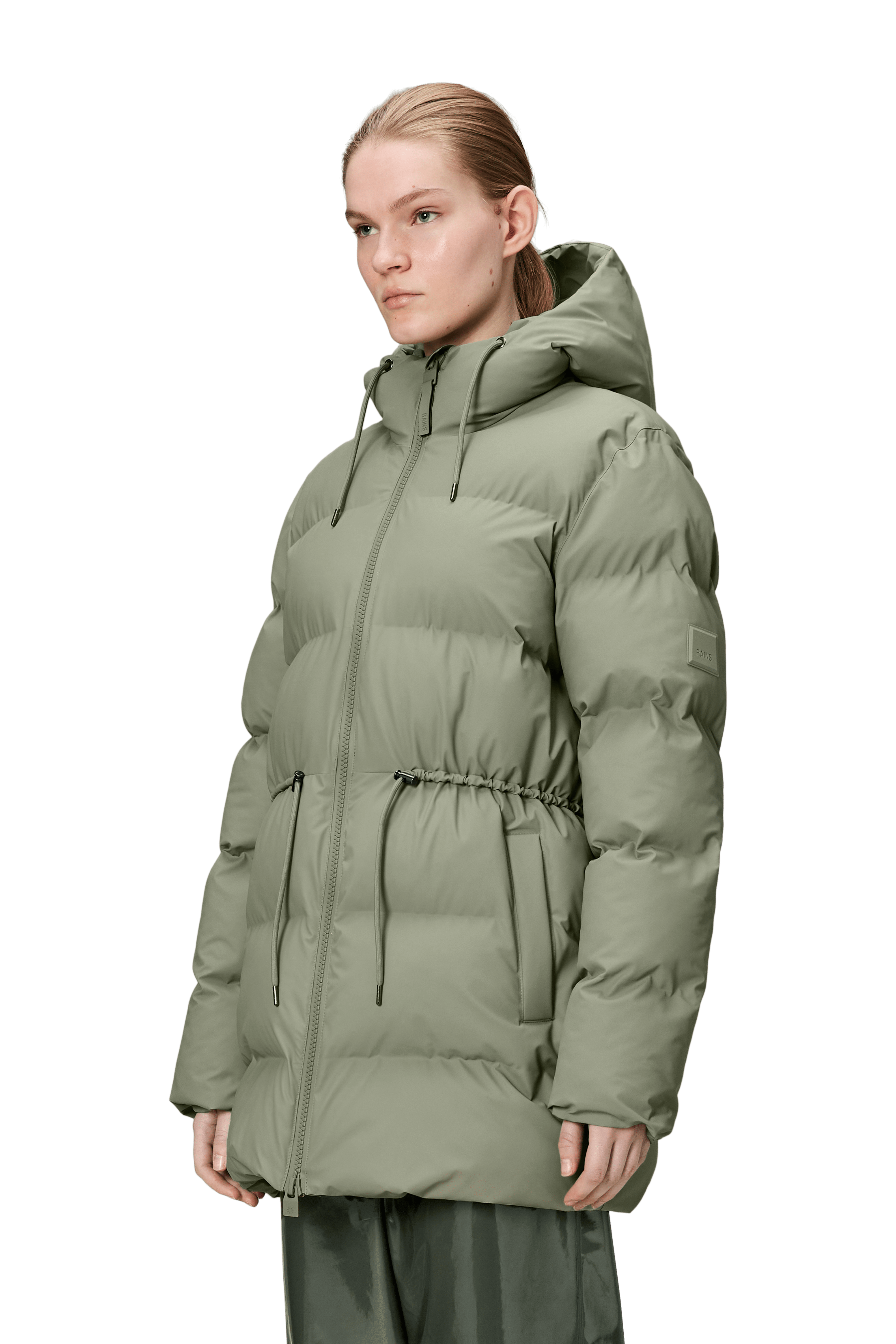 A person wearing the Alta Long Puffer String Jacket - Drift by Rains, a light olive green waterproof winter coat with an insulated hood, front zipper, drawstring waist, and side pockets, stands against a plain background.
