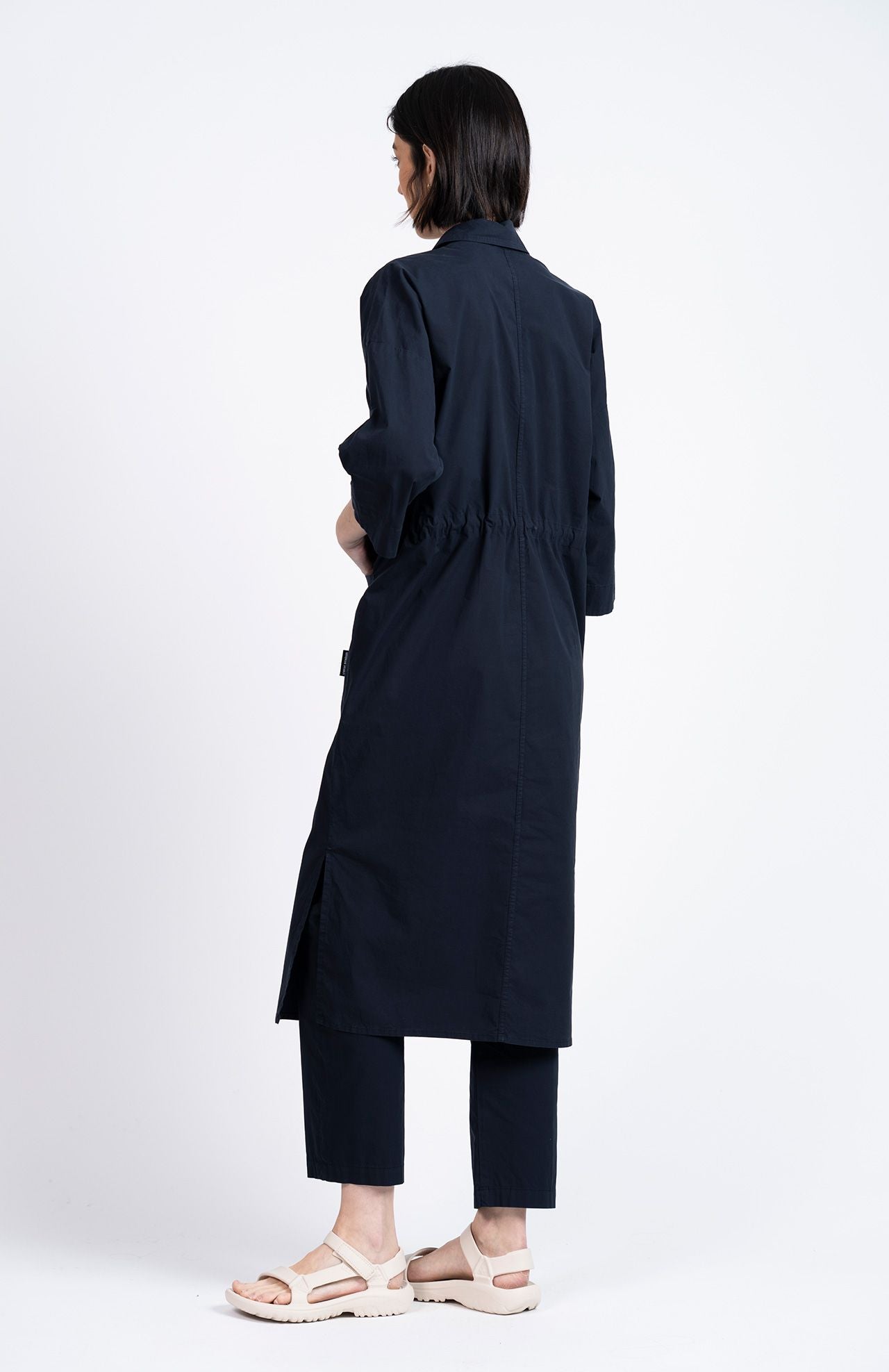 A person stands against a white background, facing away, wearing the Loreak Mendian Lore Dress in navy over dark pants with light sandals.
