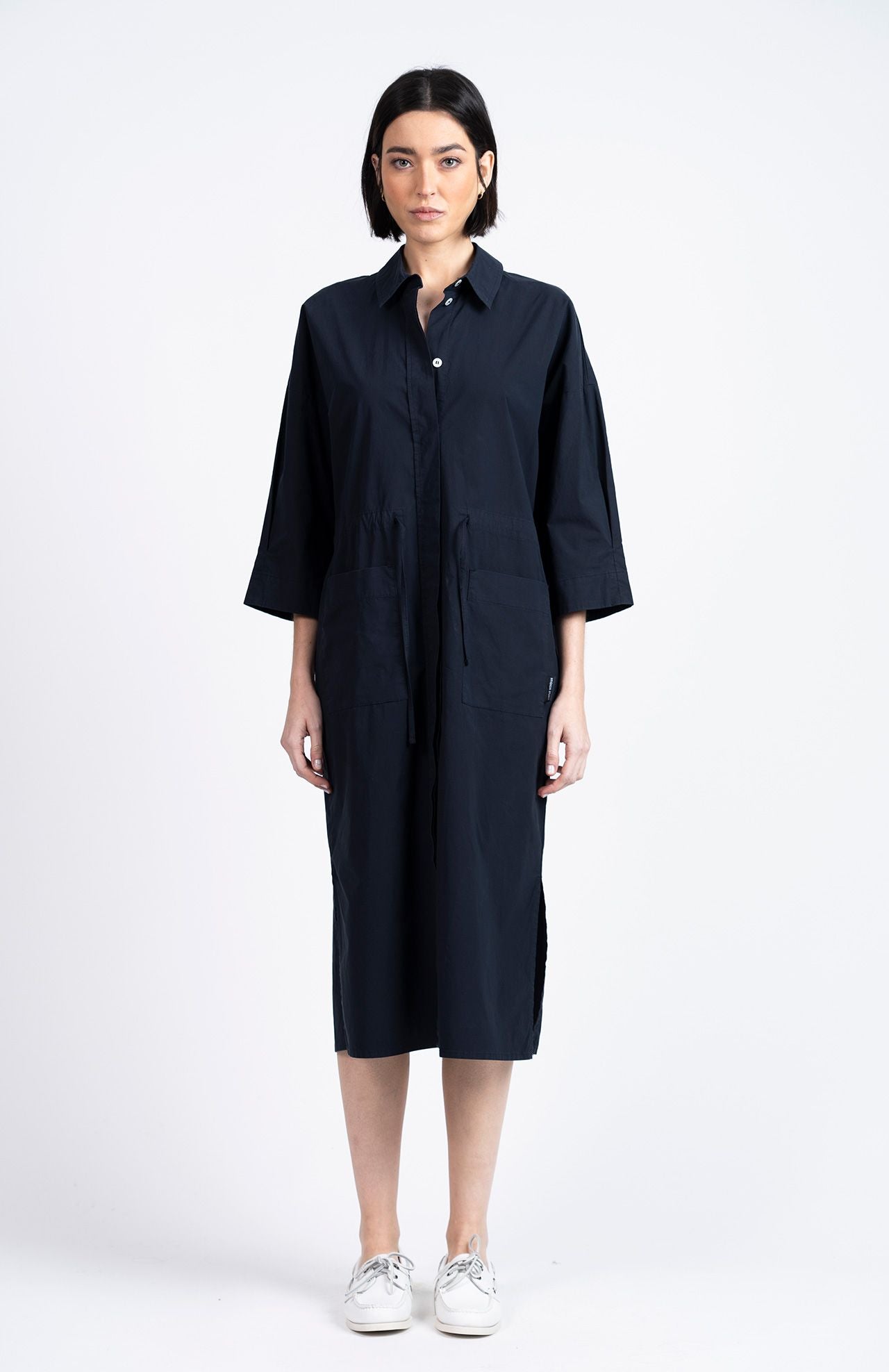 A woman faces forward wearing the Loreak Mendian Lore Dress in navy, a loose-fitting, oversized shirt dress with long sleeves, paired with white sneakers, set against a plain white background.
