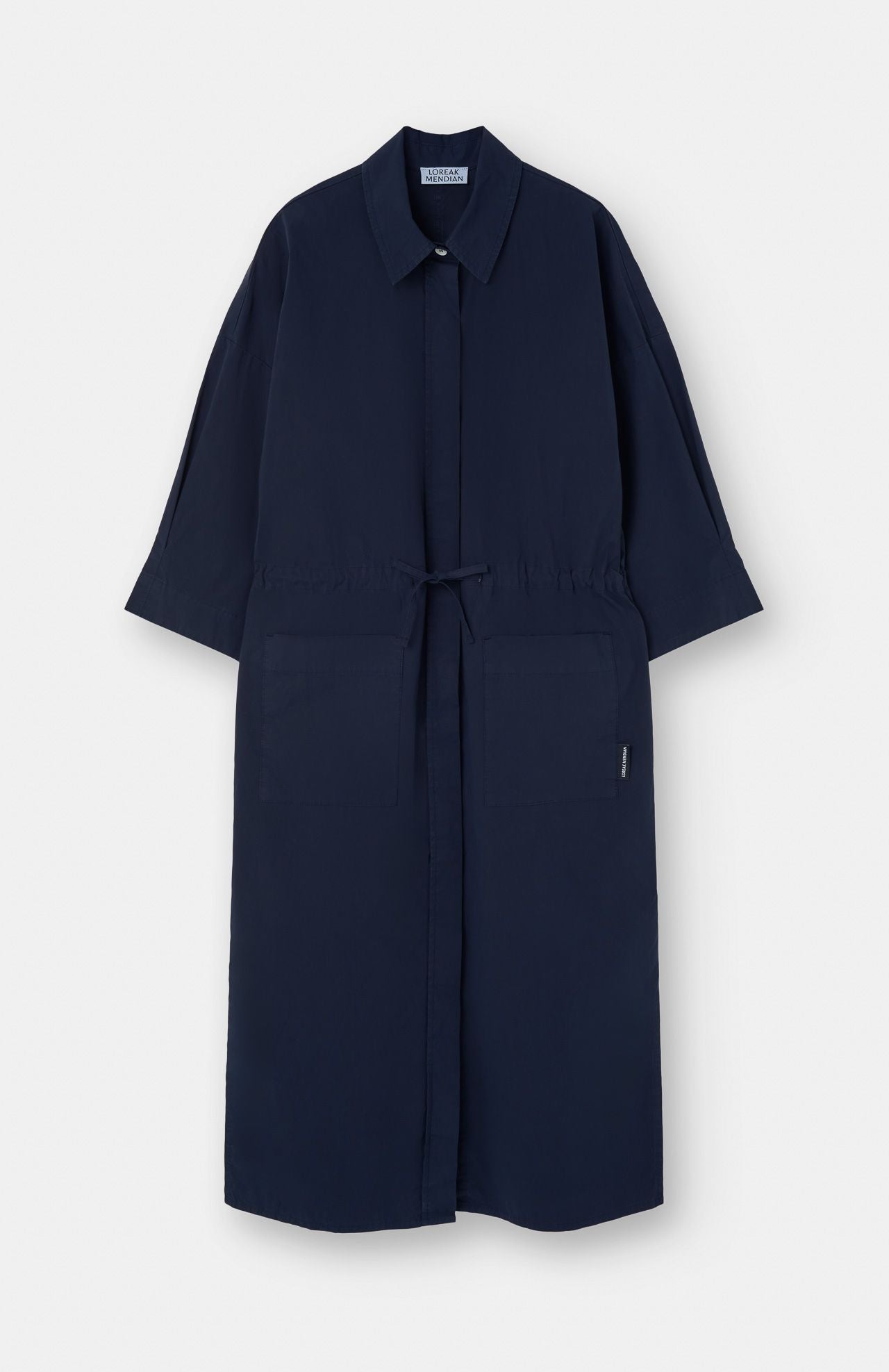 The Lore Dress in navy by Loreak Mendian is a knee-length, oversized shirt dress with a collar, front tie closure, large front pockets, and three-quarter sleeves, shown against a white background.