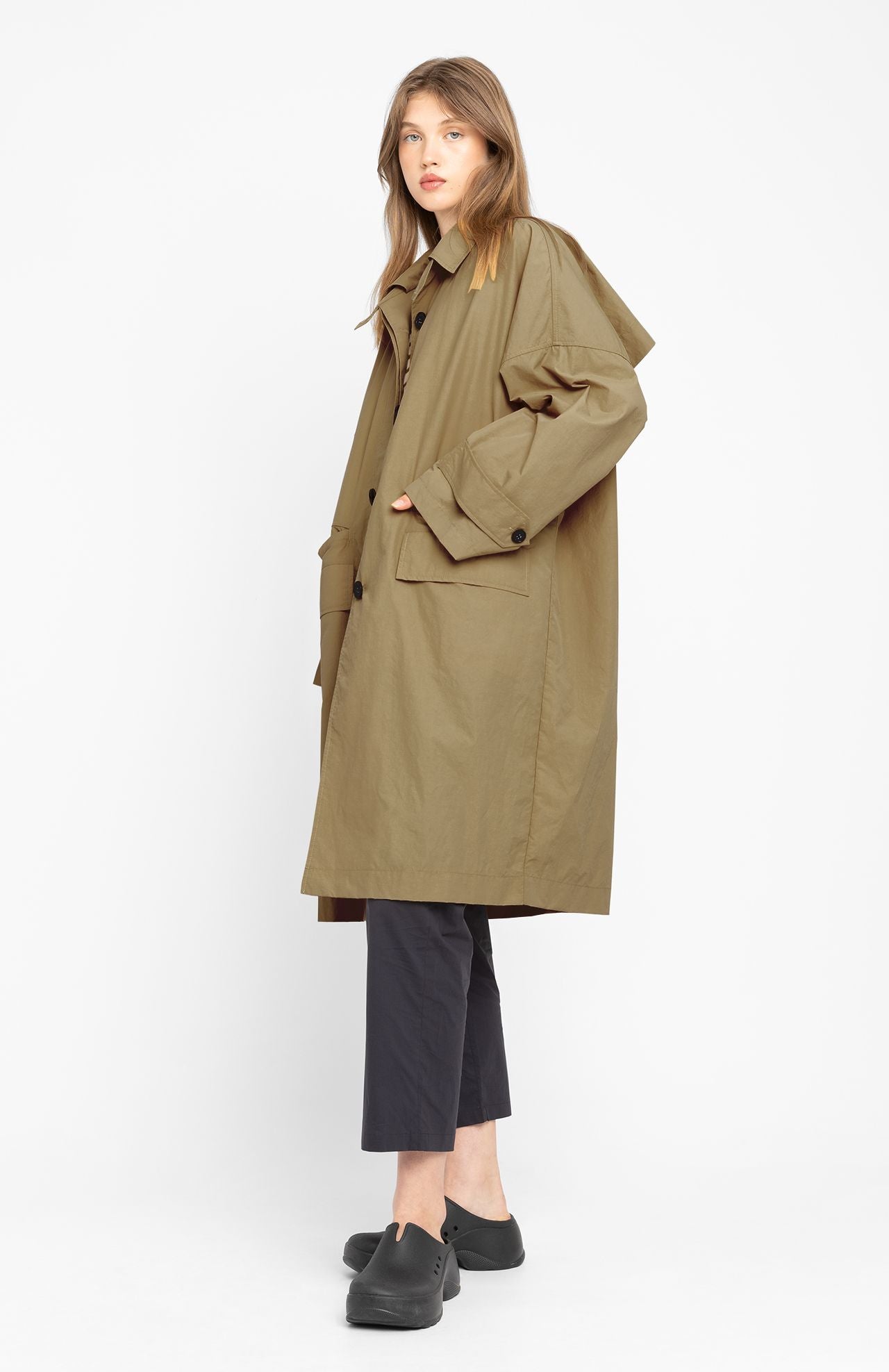 A person stands sideways against a plain white background, wearing the Loreak Mendian Lizaso Jacket in camel, paired with black pants and black slip-on shoes.