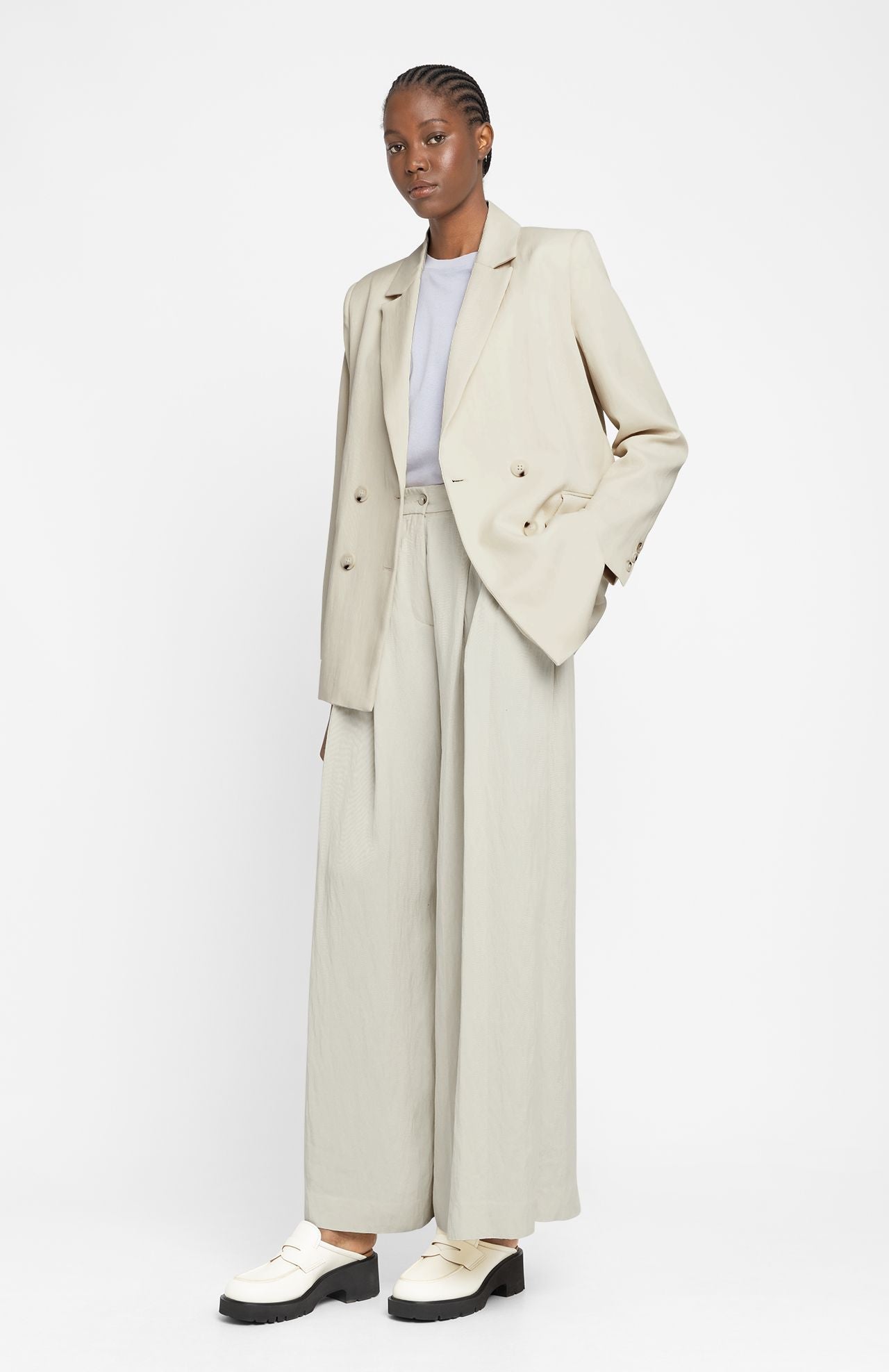 A person stands against a white background, wearing the Suarbe Tailored Jacket - Cream by Loreak Mendian in sustainable Lyocell fabric, paired with wide-leg pants, a gray shirt, and white shoes.