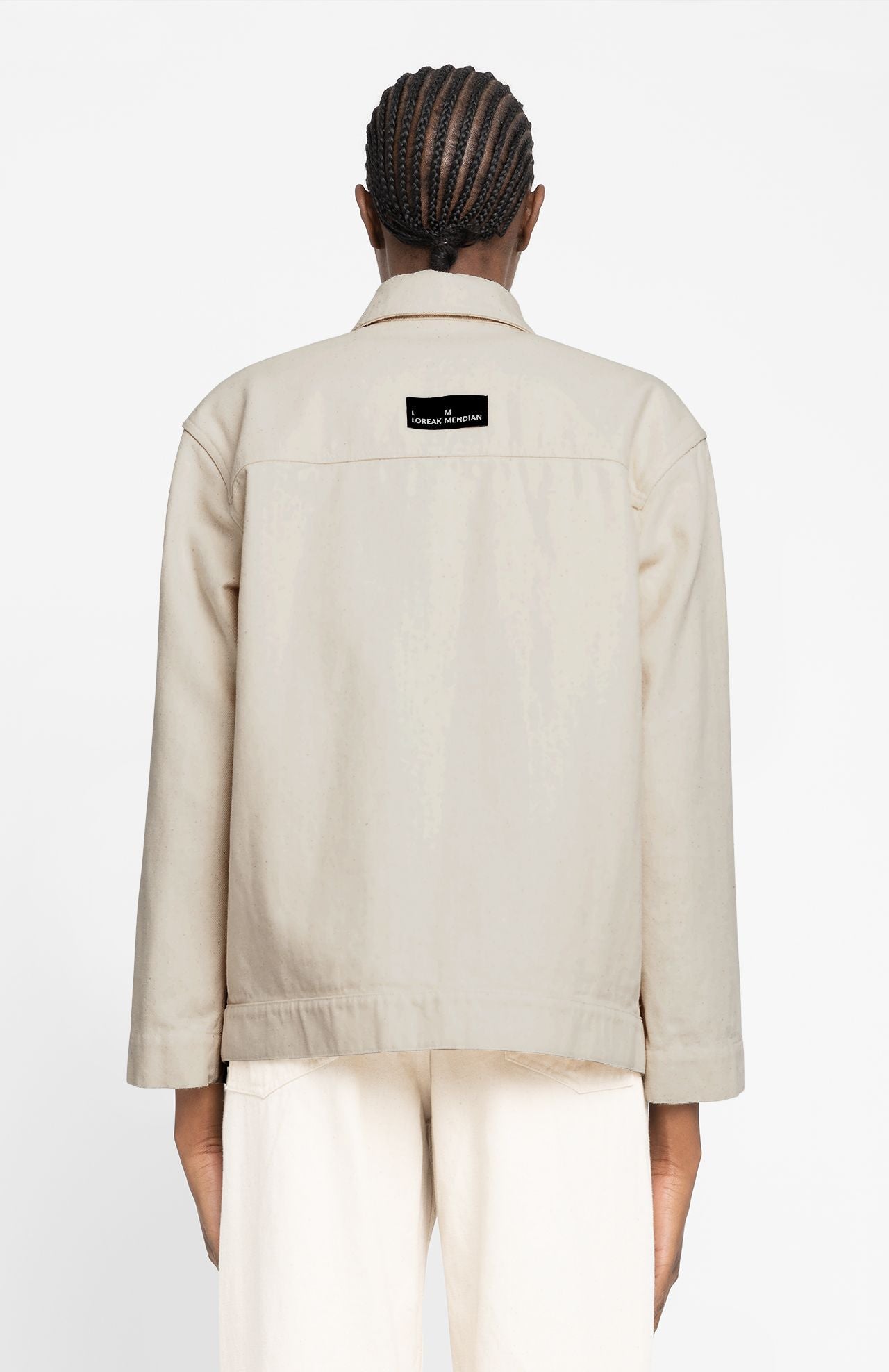 Against a plain white background, a person wearing Loreak Mendian's Igoa Jacket in ecru, featuring a black and white label on the back, pairs it with cream-colored pants while standing facing away.