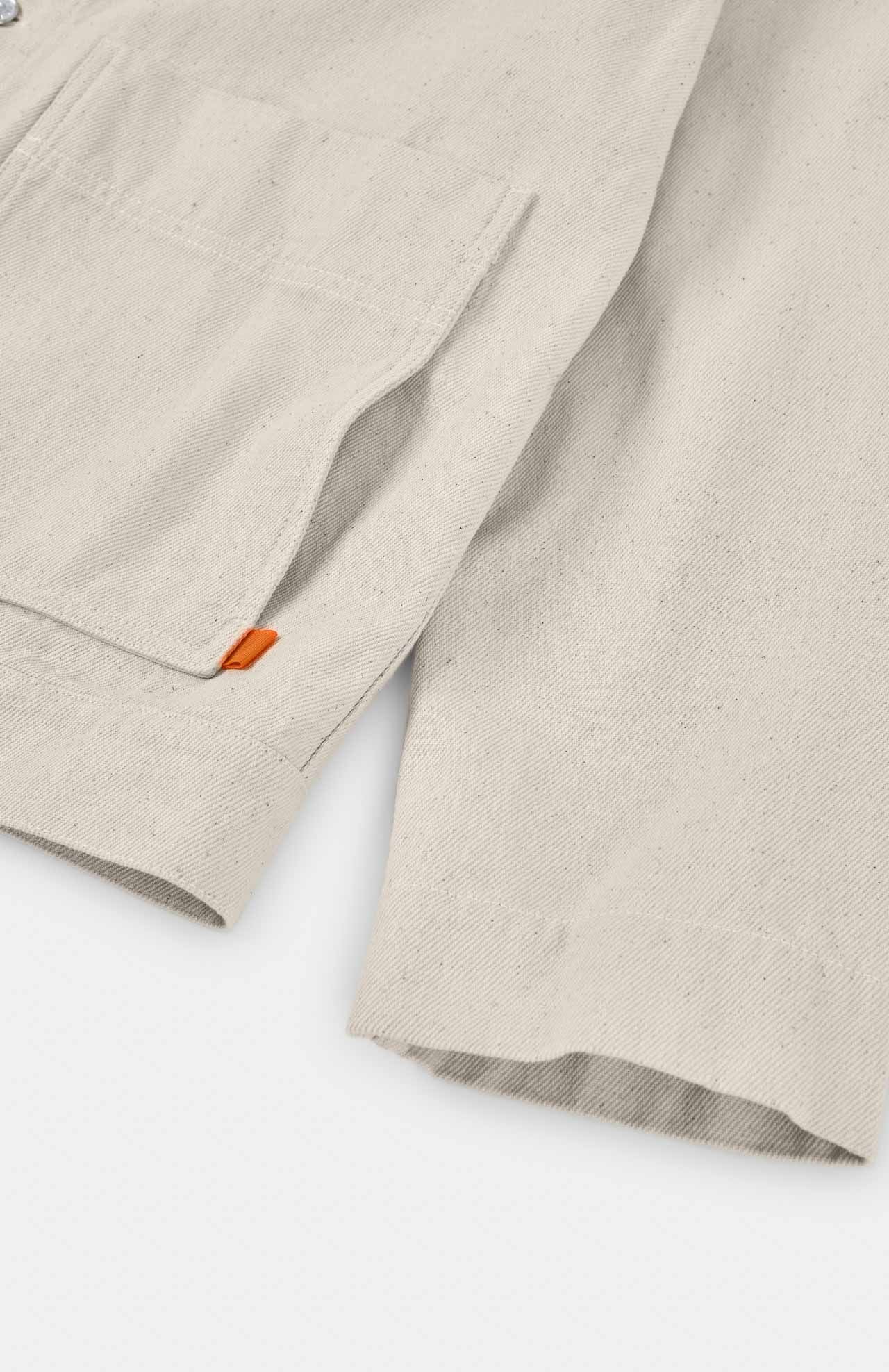 The Igoa Jacket by Loreak Mendian in ecru features a close-up of beige fabric with a left pocket and an orange tag at the seam, resembling recycled cotton and evoking a worker-style garment.