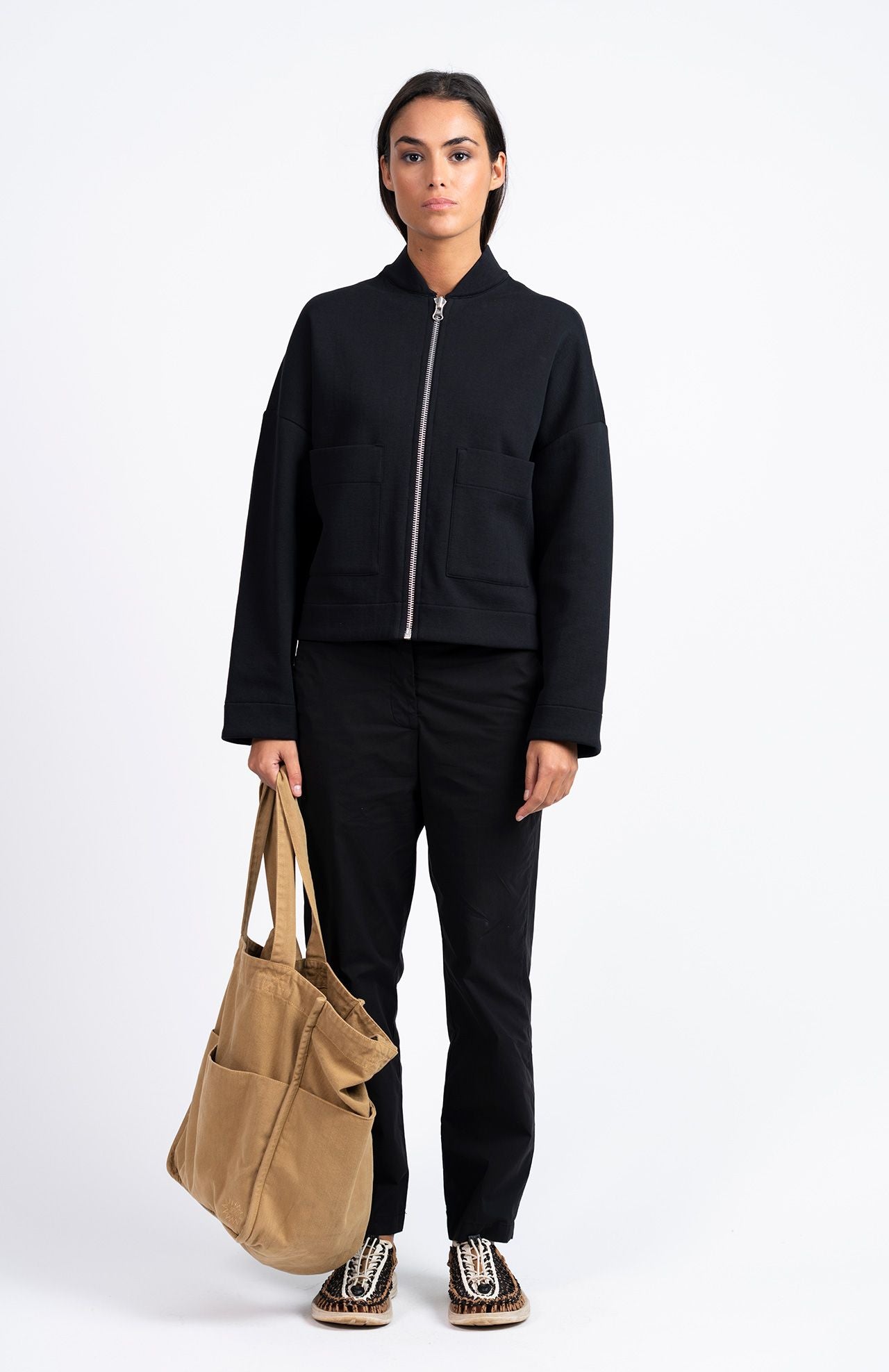 A person stands facing forward against a plain white background, wearing the Loreak Mendian Lili Cropped Jacket in black with black pants, patterned shoes, and holding a tan tote bag.