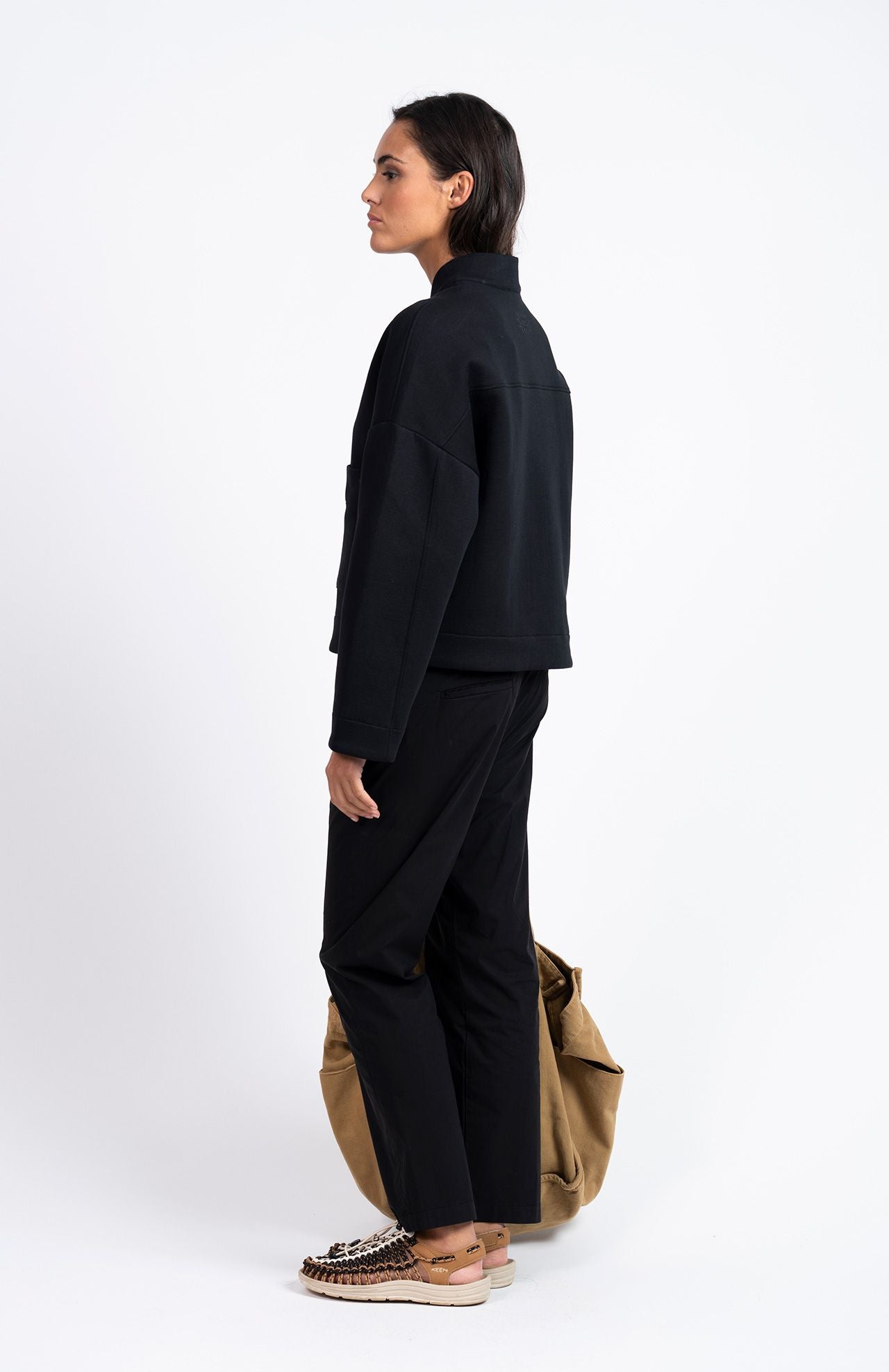 A woman stands in profile against a white background, wearing the Loreak Mendian Lili Cropped Jacket in black, paired with black pants, printed slip-on shoes, and holding a large tan bag by her side.