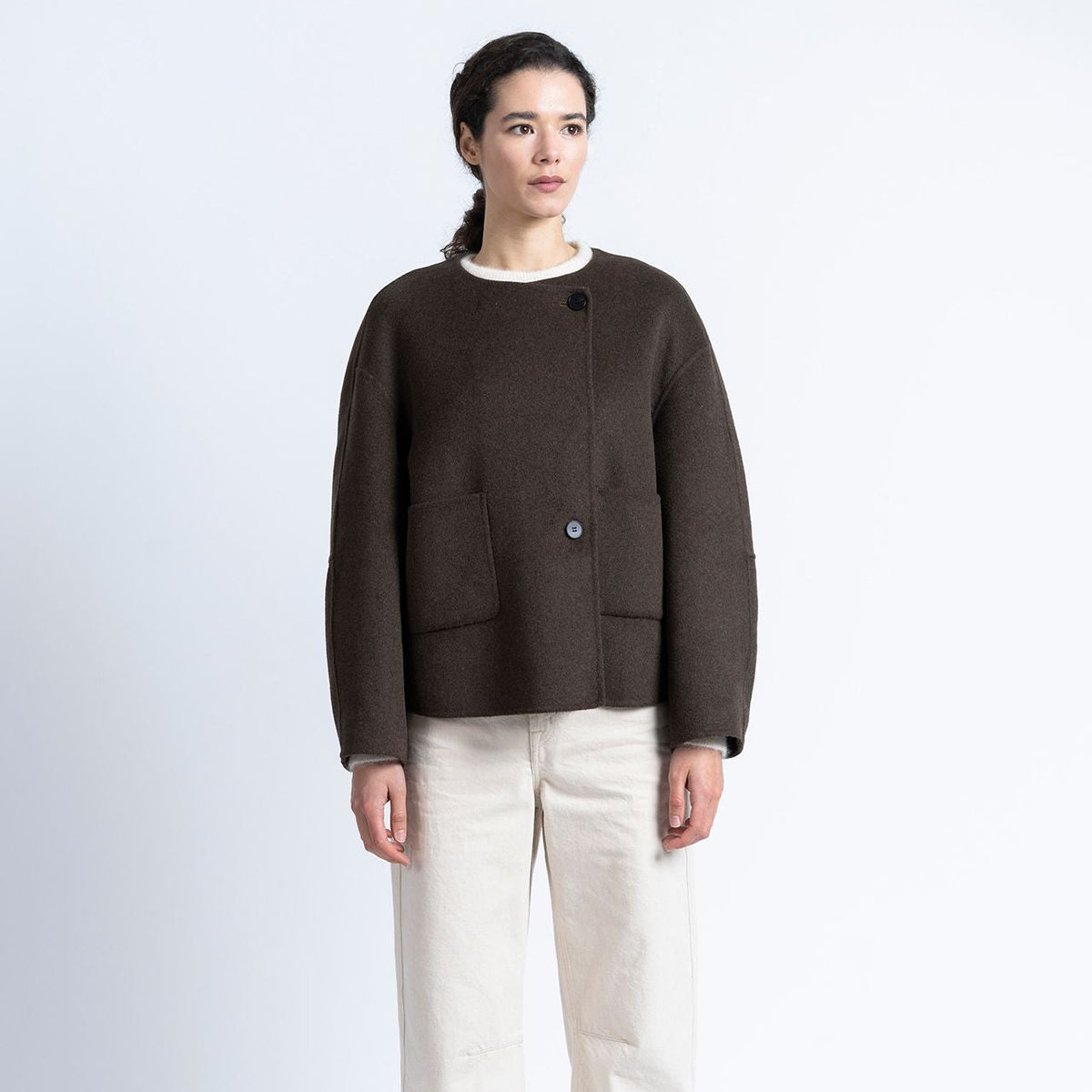 A model stands against a light gray studio background wearing a dark brown cropped button-front jacket with front patch pockets, paired with off-white wide-leg trousers and dark brown shoes.