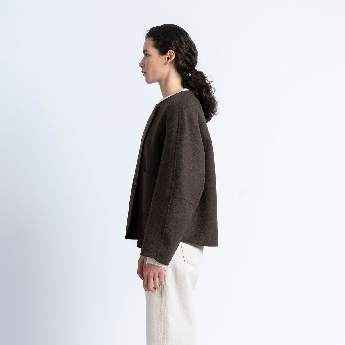 A woman shown in profile wearing a dark brown cropped jacket, off-white wide-leg trousers, and black shoes, standing against a plain light gray studio background.
