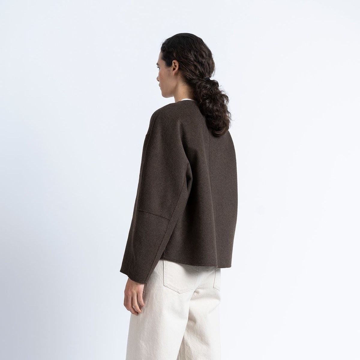 A model shown from the back wearing a dark brown cropped knit sweater, off-white wide-leg trousers, and dark brown shoes, standing against a plain white studio background.