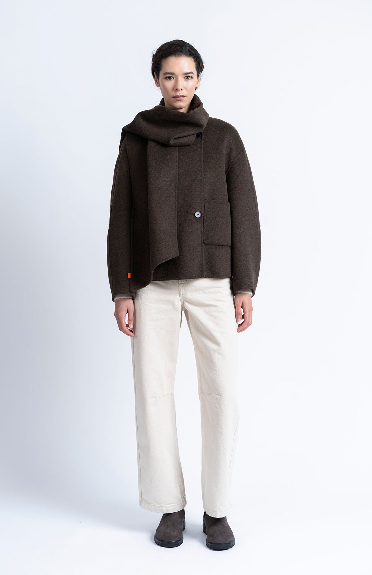 A model stands against a light gray studio background wearing a dark brown asymmetrical wrap jacket, a matching dark brown scarf, off-white wide-leg trousers, and dark brown shoes.