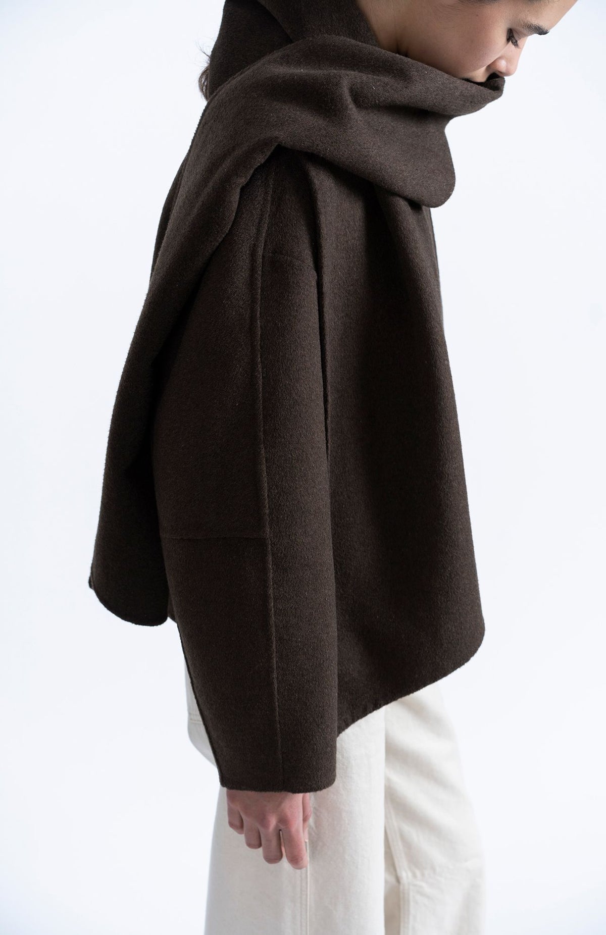 A side profile view of a person wearing a dark brown draped cape-style jacket with a high folded collar, paired with off-white trousers, against a plain white background.