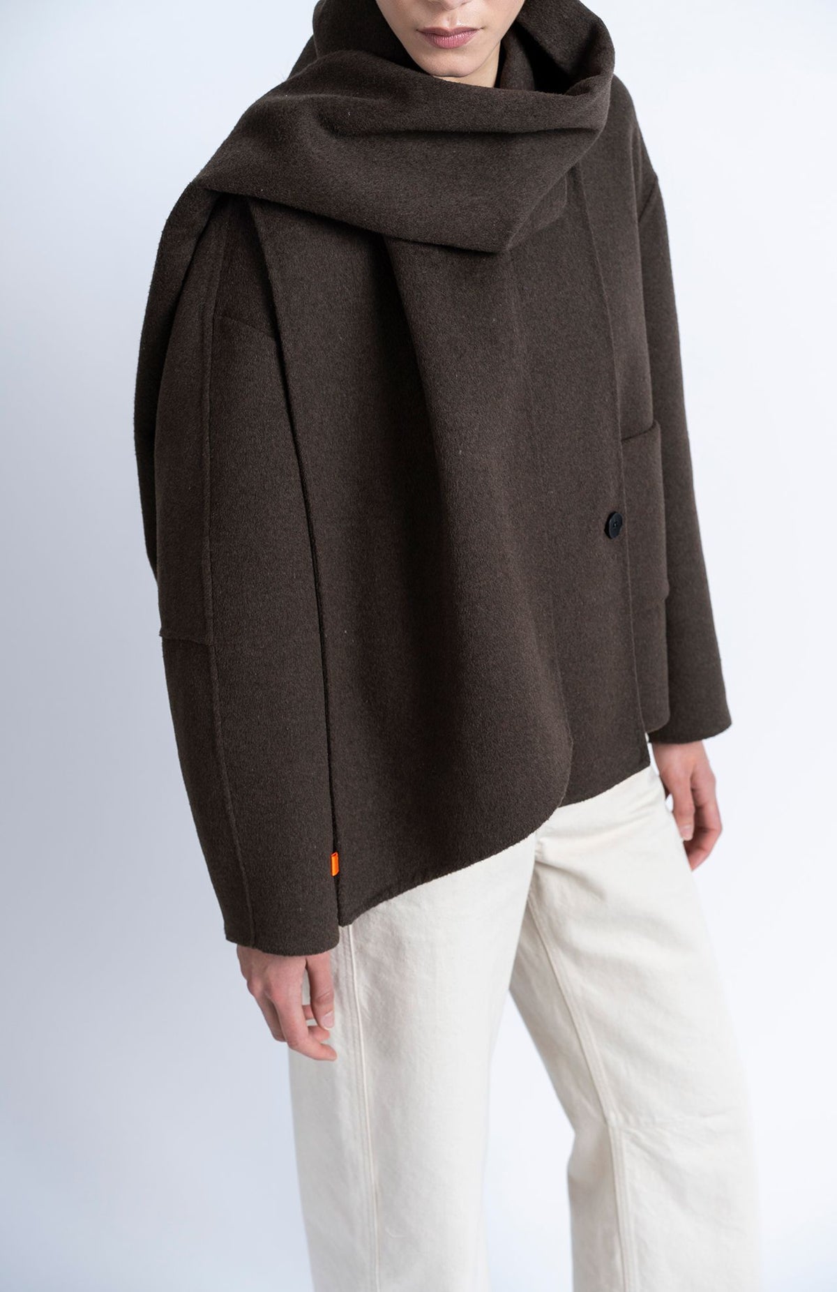 A model wearing a dark brown oversized draped jacket with a single button closure and cream-colored trousers, photographed against a light gray studio background.