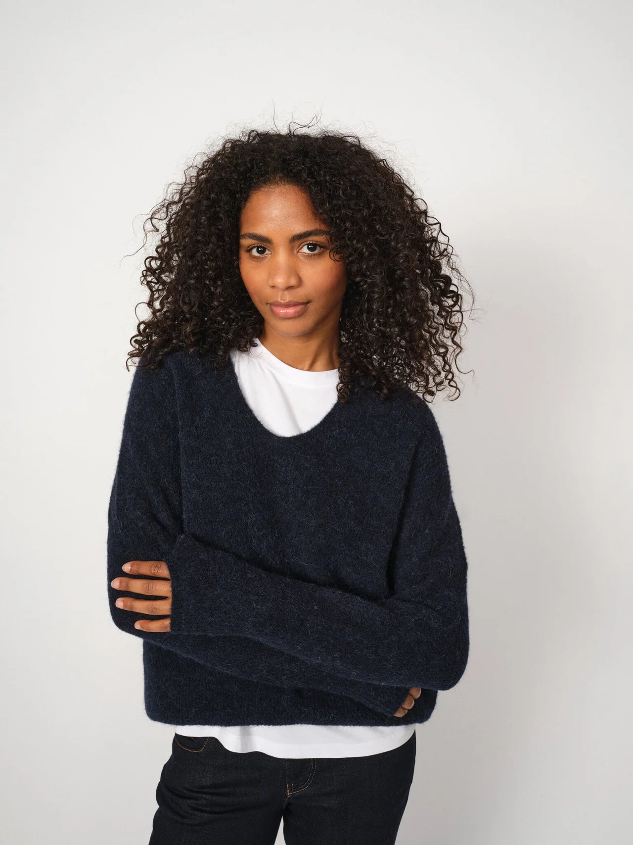 A person with curly hair stands against a plain white background, arms loosely crossed, wearing the MOS MOSH Thora V-Neck in Navy— a relaxed fit women's sweater crafted from soft Alpaca Wool and layered over a white shirt.