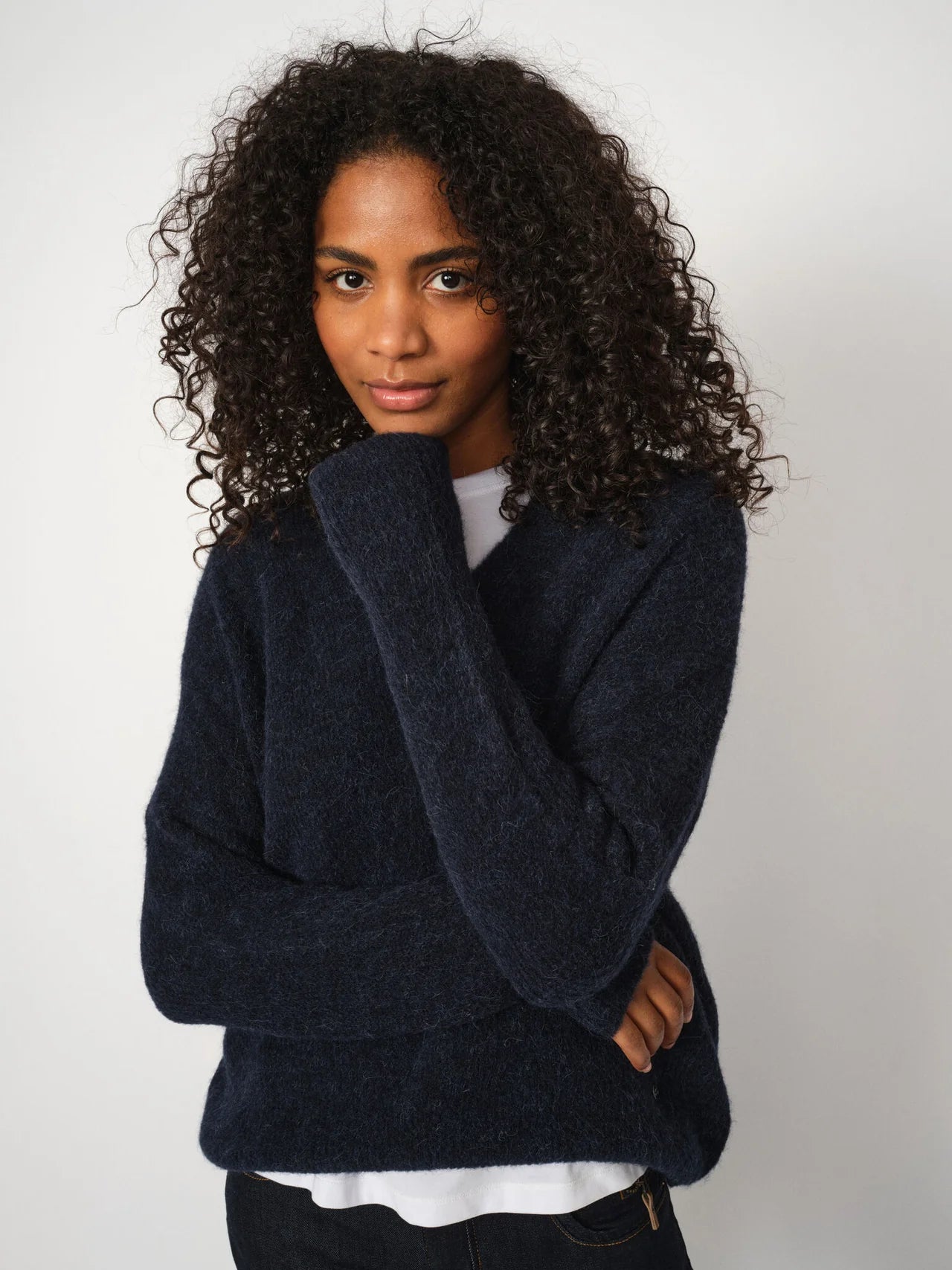 A person with curly hair wears the MOS MOSH Thora V-Neck sweater in navy over a white shirt, standing against a plain white background and looking at the camera with a neutral expression.