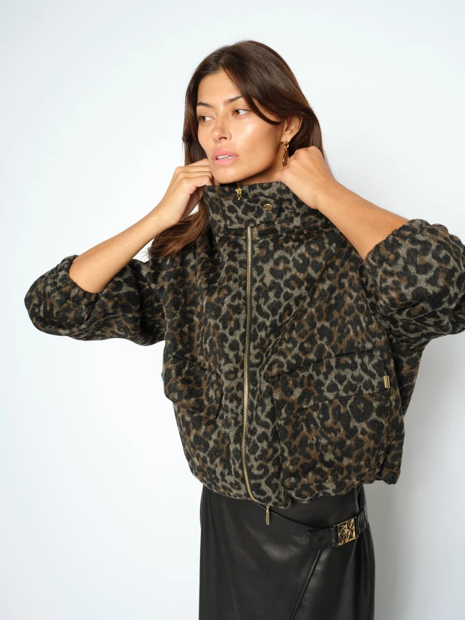 A woman zips up the MOS MOSH Camille Leo Jacket in black—an oversized, leopard-print style with a high collar—paired with a black skirt featuring a decorative buckle, standing against a plain background.