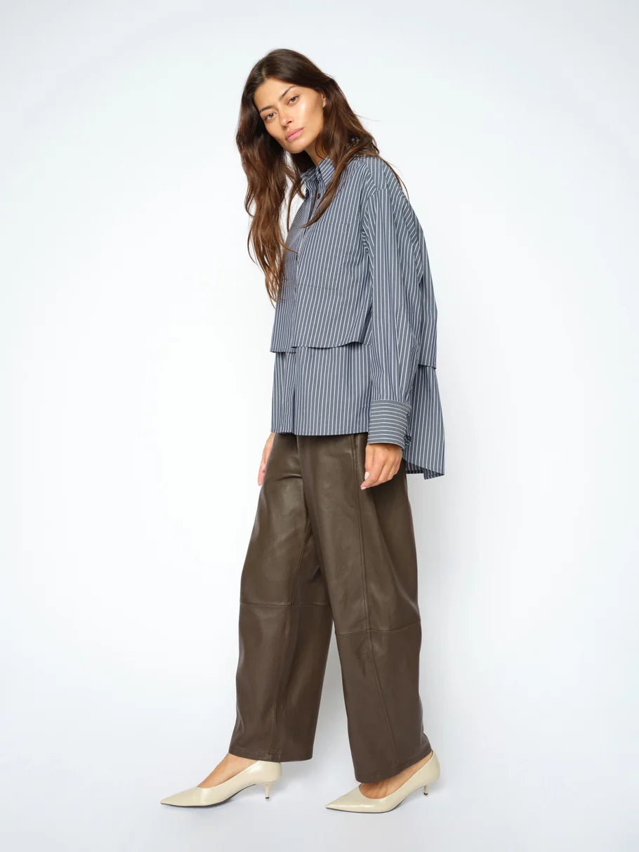A woman wearing the MOS MOSH Barrel Leather Pant in wide brown lamb leather with a barrel-leg silhouette, paired with a blue striped shirt and cream heels, stands against a plain light background.