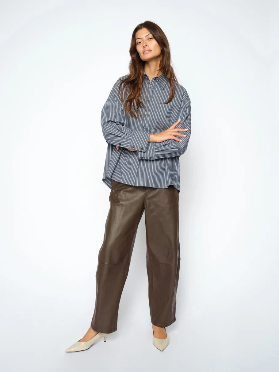 A woman stands against a white background wearing a blue striped shirt, MOS MOSH Barrel Leather Pant in brown lamb leather with a barrel-leg silhouette, and beige pointed-toe shoes, her arms crossed and head tilted slightly.
