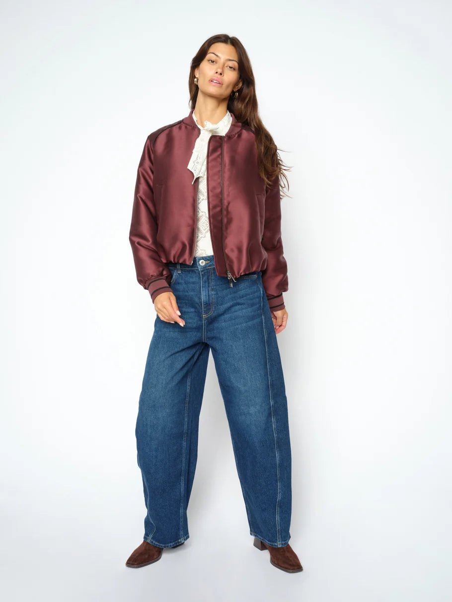 A woman stands against a white background wearing a maroon satin bomber jacket, white blouse, brown shoes, and the MOS MOSH Cindy Draciel Jeans—a pair of high-rise organic cotton jeans with a wide-leg silhouette.