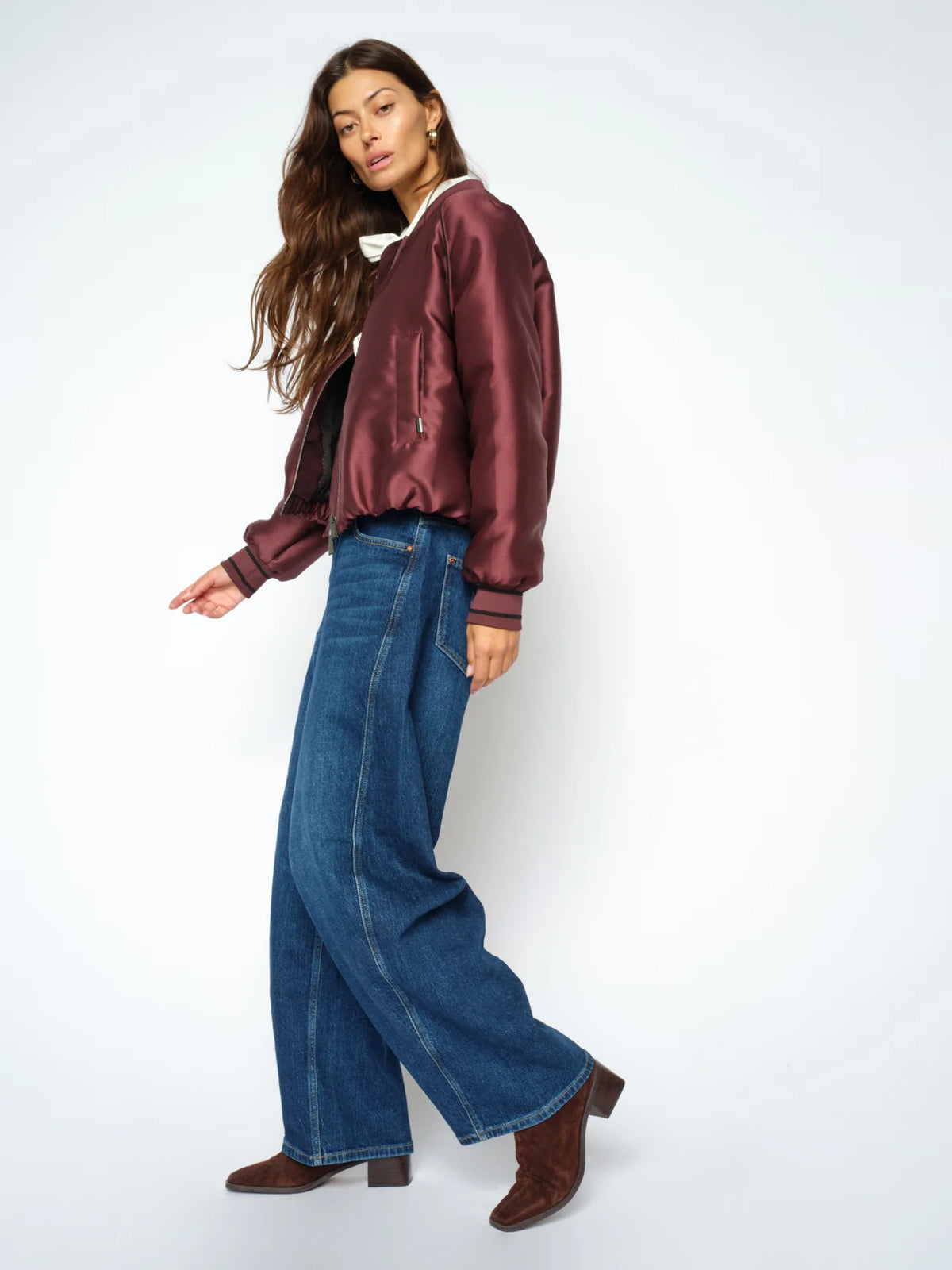Wearing MOS MOSH Cindy Draciel Jeans—high-rise barrel-leg jeans made from organic cotton—a woman poses in a maroon satin bomber jacket and brown heeled boots against a plain white background.