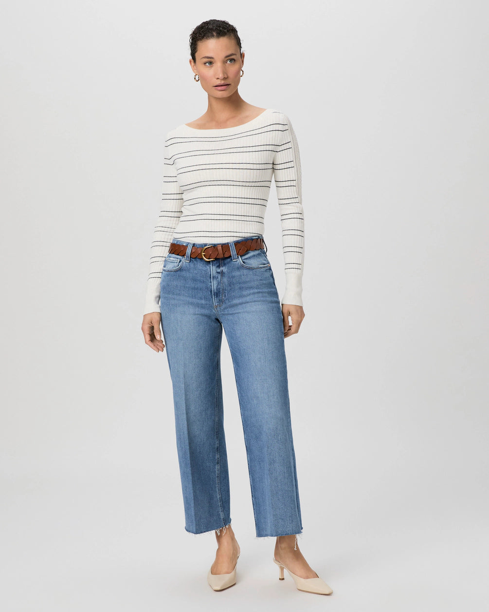 A person wearing a white striped sweater, Paige's Anessa Wide Leg - Poet ultra high-waisted blue jeans, a brown belt, and beige heels stands against a plain background.