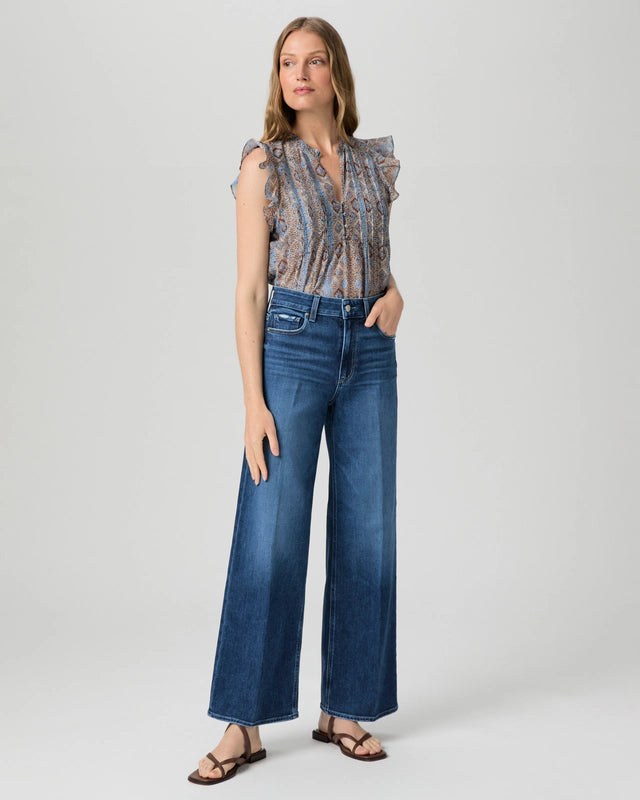 A woman stands against a plain background wearing a sleeveless patterned blouse, brown sandals, and Paige’s Anessa Wide Leg 31"- Dresden jeans.