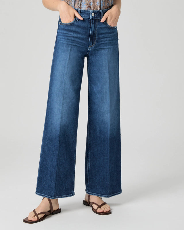 A person wears Paige’s Anessa Wide Leg 31"- Dresden denim jeans in vintage blue with brown sandals, hands in pockets, shown from shoulders down against a plain background.