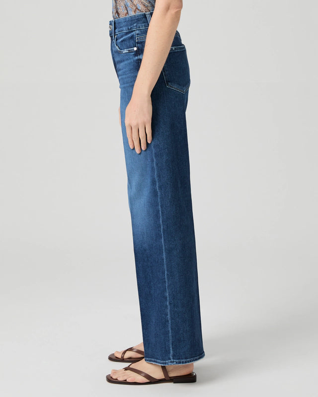 A person in profile wears Paige's Anessa Wide Leg 31"- Dresden jeans, a patterned top, and brown sandals against a plain background.