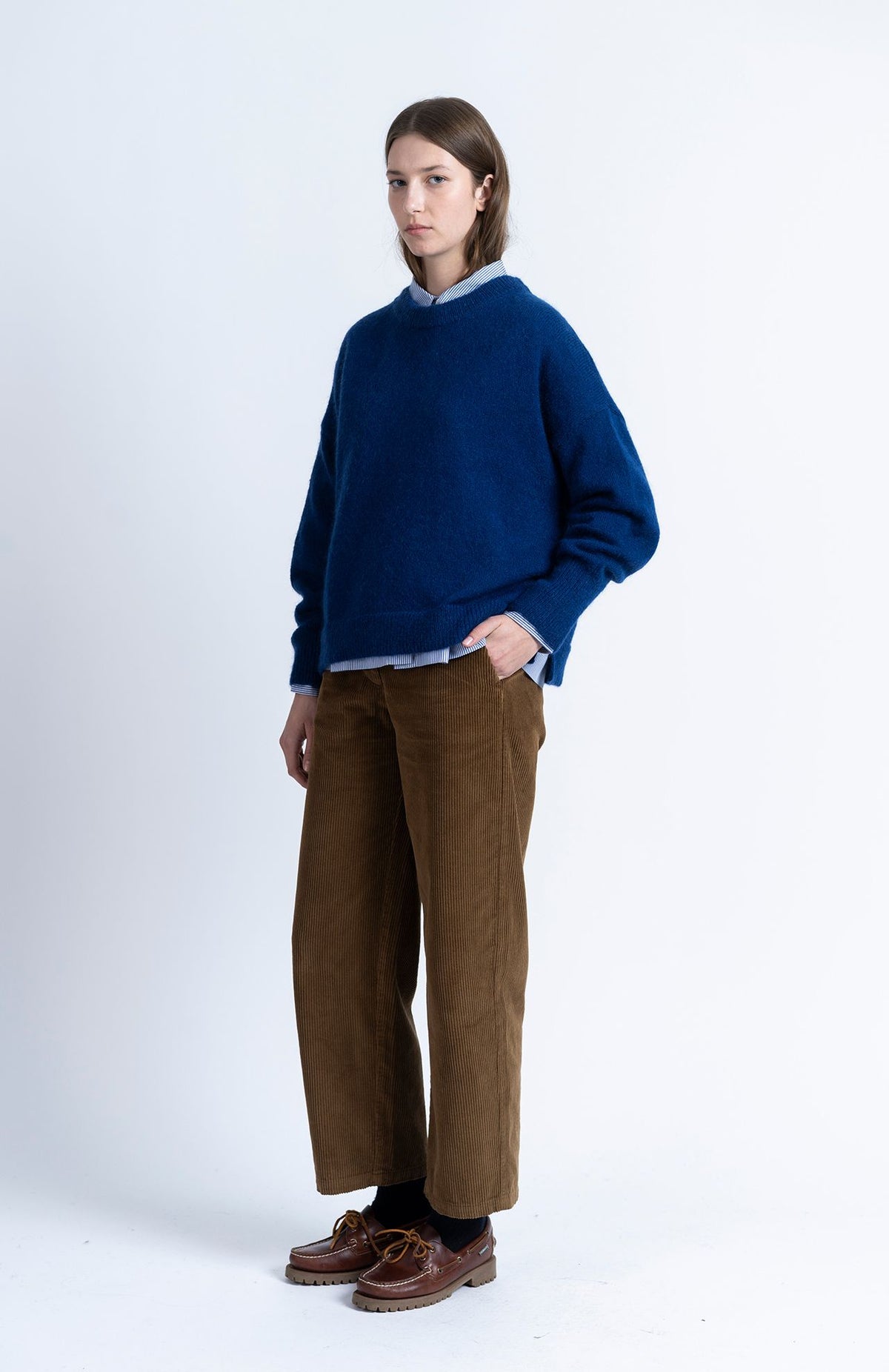 A person wearing a navy Loreak Mendian Geroa Jumper over a striped button-down shirt, brown corduroy trousers, and brown leather boat shoes.