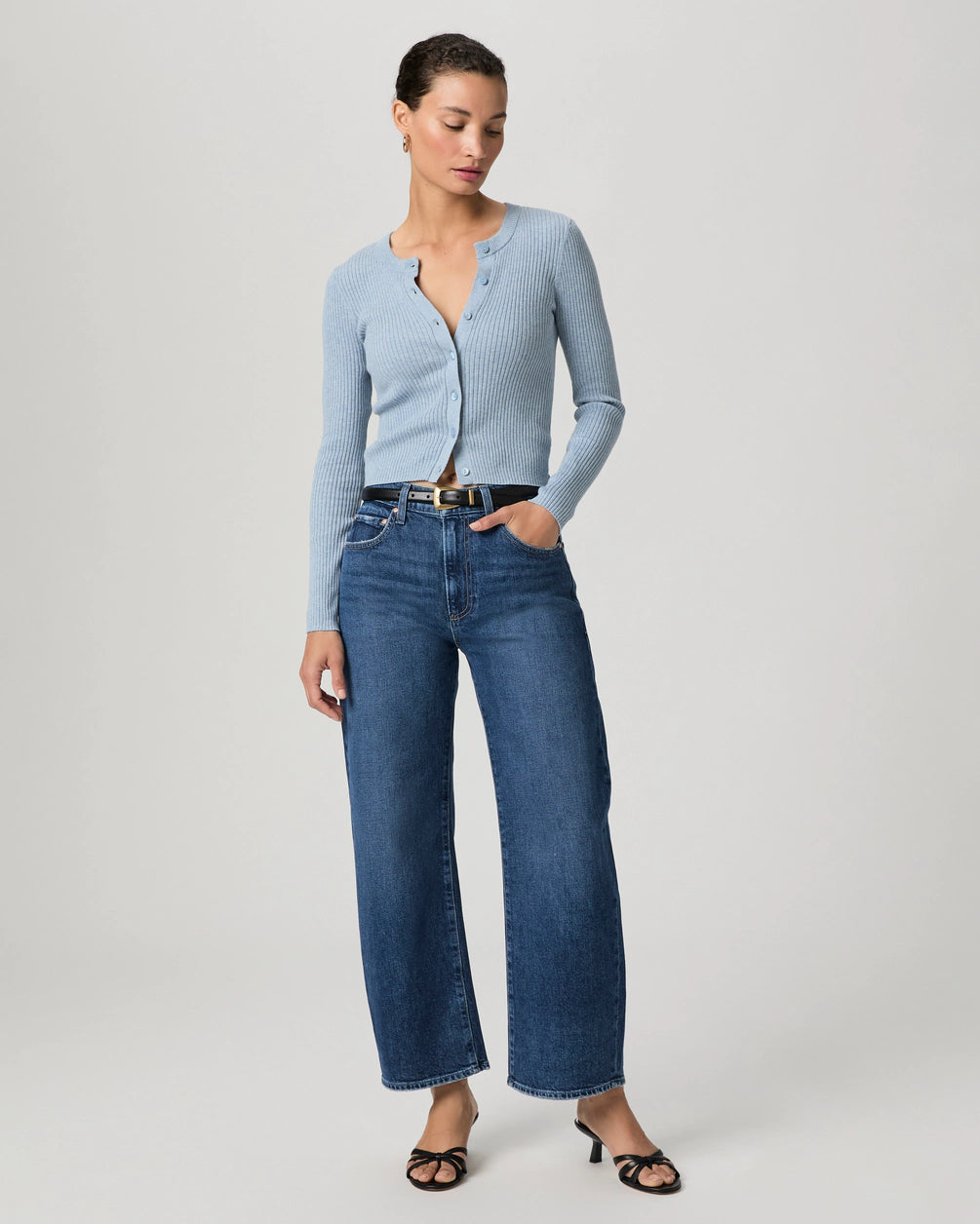 A woman stands against a plain background wearing a light blue ribbed cardigan, Paige Arellia Ankle Barrel Leg - Soundcheck jeans, a black belt, and black heeled sandals.