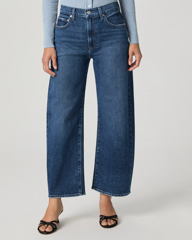 Wearing Paige's Arellia Ankle Barrel Leg jeans in Soundcheck, styled with black heeled sandals and a light blue ribbed long-sleeve top, standing against a plain background.