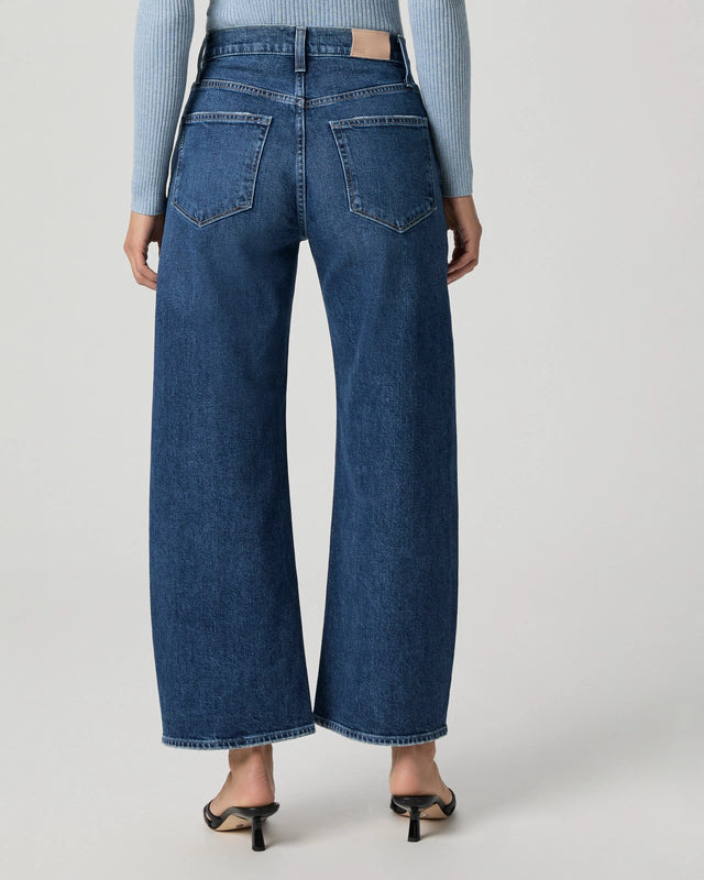 A person is shown from the back against a plain light background, wearing Paige Arellia Ankle Barrel Leg - Soundcheck jeans in a vintage wash with black heeled sandals.