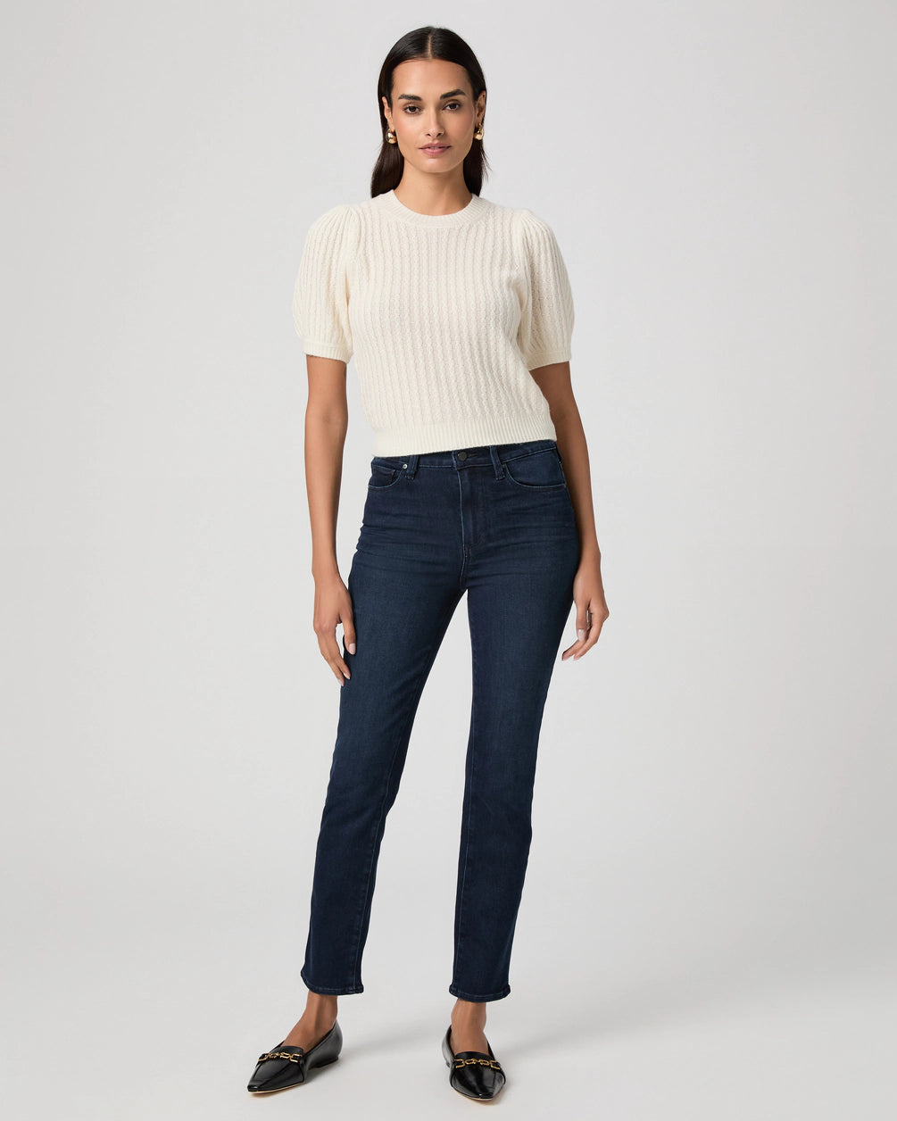 A woman stands against a plain background wearing a white short-sleeve knit sweater, Paige Gemma Cigarette Leg - Bespoke jeans, and black flats.
