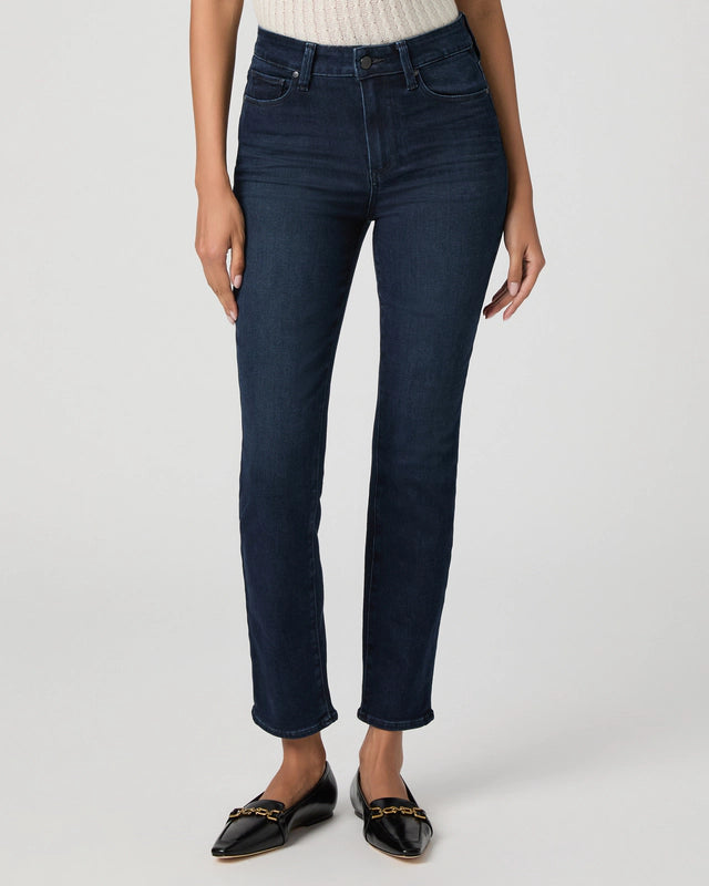 A person wearing Paige Gemma Cigarette Leg - Bespoke jeans, a light ribbed top, and black pointed-toe flats with a gold chain detail stands against a plain light background.