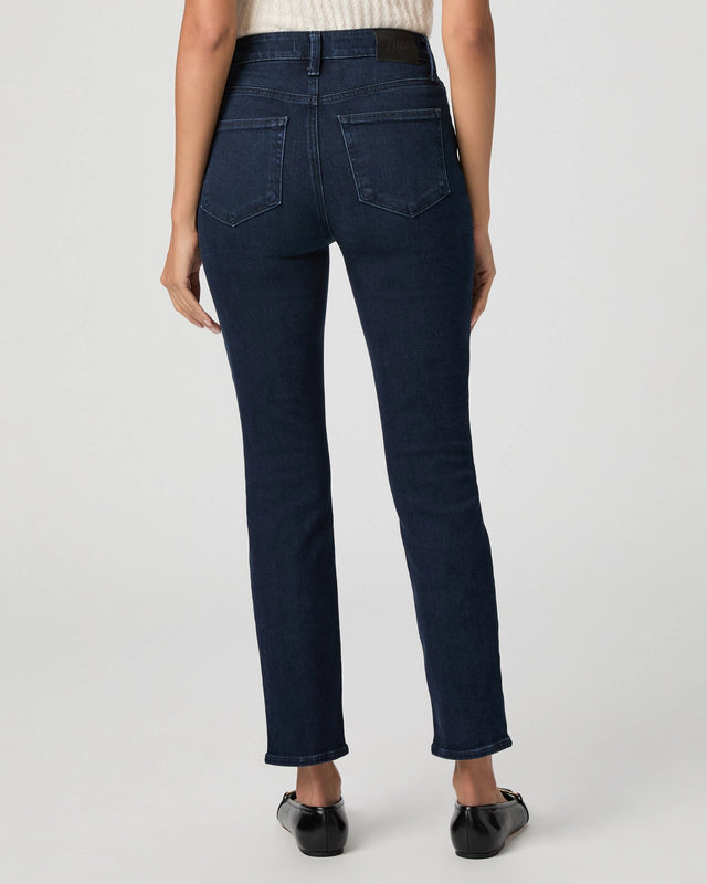 A person stands facing away, wearing Paige Gemma Cigarette Leg - Bespoke jeans in dark blue with an ankle-length inseam, a light top, and black flat shoes against a plain background.