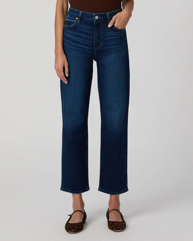 A person wearing Paige Mason Ankle Straight - Overdrive denim high-waisted, dark blue straight-leg jeans, a dark top, and leopard print flats stands against a plain light background.