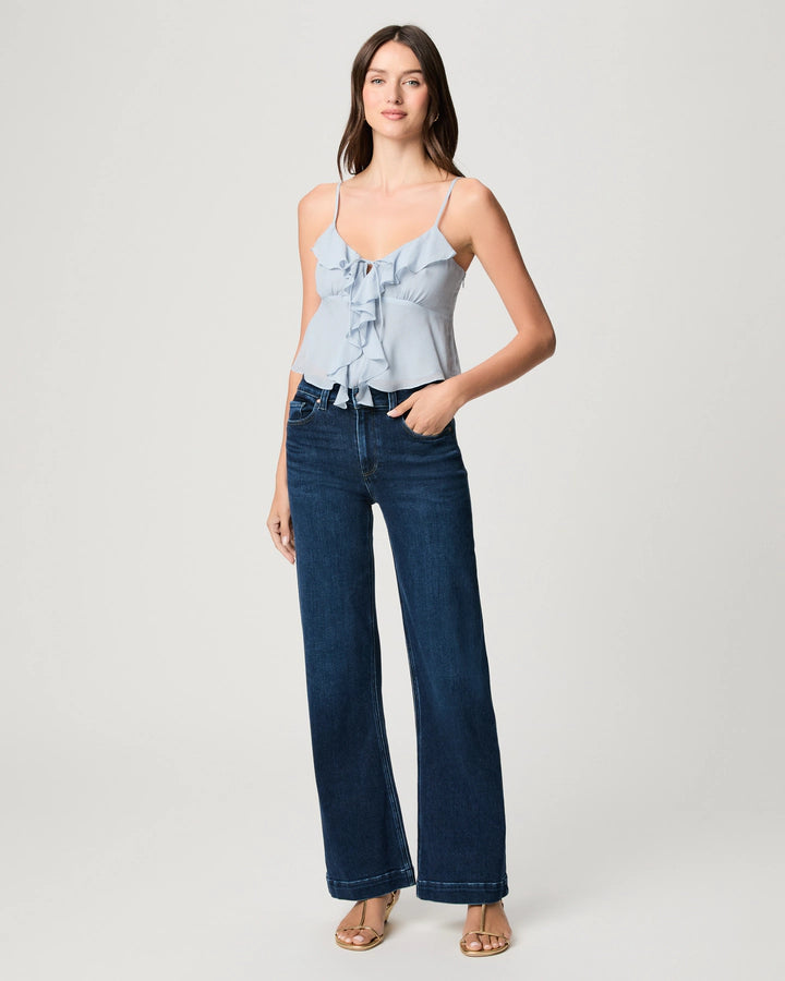 A woman stands in a studio wearing a light blue ruffle camisole, Paige Leenah Slim Wide Leg jeans in Basilica, and sandals, with one hand in her pocket.