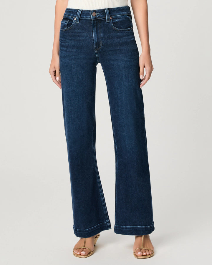 A person is wearing the Paige Leenah Slim Wide Leg jeans in vintage Basilica wash, featuring a high-rise, wide-leg fit with front pockets, paired with brown sandals and standing against a plain light background.