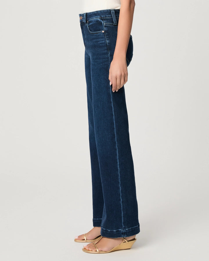 A person wearing the Paige Leenah Slim Wide Leg jean in the classic Basilica vintage wash, styled with a white ribbed tank top and beige open-toe sandals, is shown in side profile against a plain light background.