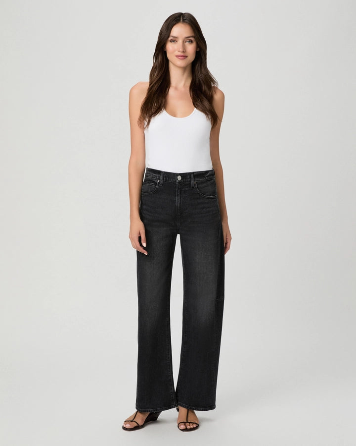 A woman stands against a plain background, facing forward with her arms relaxed at her sides, wearing a white tank top, black sandals, and the Paige Arellia Skimmer Barrel Leg jeans in Oscura—a high-rise barrel leg style in a vintage black wash that captures effortless style and the timeless appeal of Heritage denim.