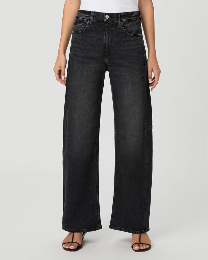 A person wearing Paige Arellia Skimmer Barrel Leg jeans in Oscura, paired with a white top and black sandals, stands against a plain light background.