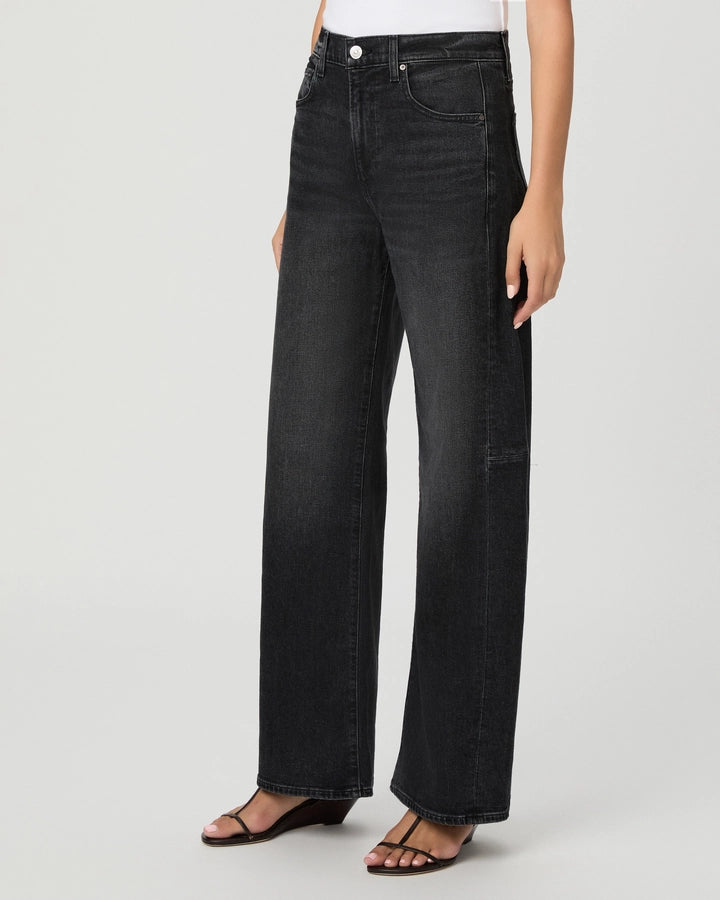 A person wearing the Paige Arellia Skimmer Barrel Leg jeans in Oscura, a vintage black wash, paired with black sandals stands against a plain light background.