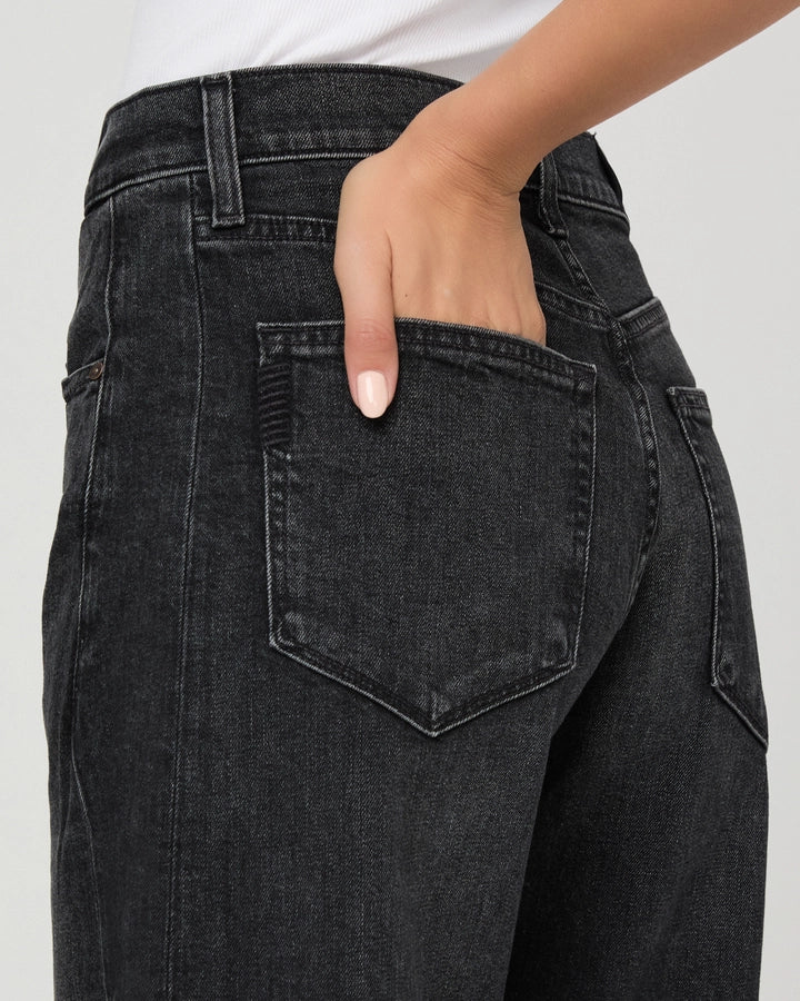 A person wearing the Paige Arellia Skimmer Barrel Leg - Oscura jeans, styled with a white top, stands against a plain background with their right hand placed in the back pocket of the jeans.